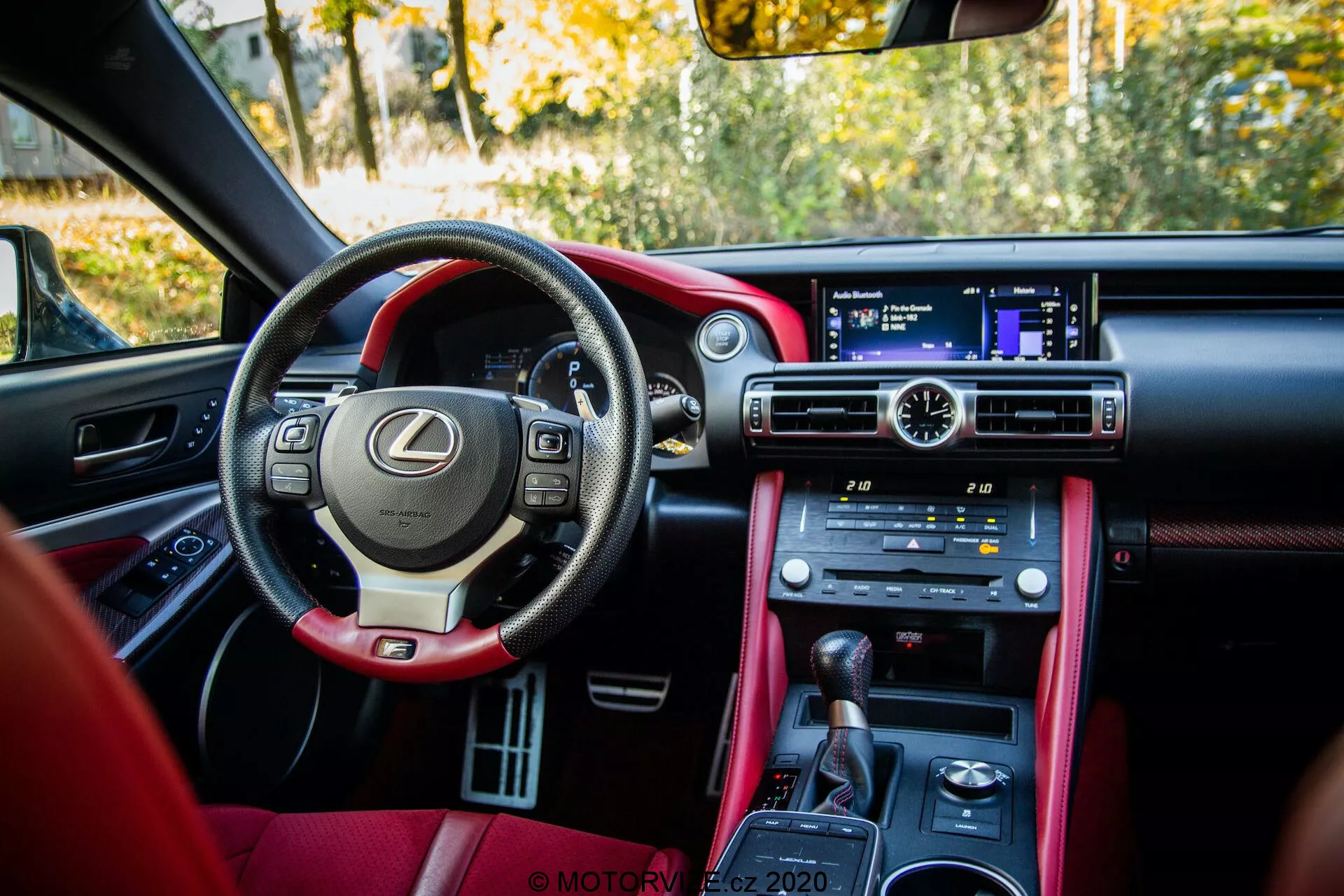 Vue intérieure d'une Lexus RC 2016 stationnée depuis l'arrière du côté passager. La prise de vue détaille un habitacle avant luxueux avec un volant en cuir équipé de commandes multifonctions et de palettes de changement de vitesse, suggérant un caractère sportif. Le tableau de bord large abrite un écran d'infodivertissement non tactile avec un pavé tactile à distance sur la console centrale gainée de cuir. Des boutons physiques pour le contrôle climatique et le chauffage/refroidissement des sièges, ainsi que des sélections de modes de conduite comme Eco, Normal et Sport, sont visibles. L'intérieur bicolore, en cuir foncé et rouge, est baigné par la lumière naturelle, montrant un environnement extérieur stationnaire avec feuillage suggérant une scène automnale.