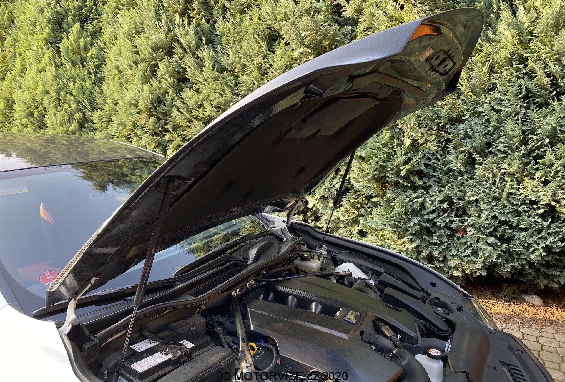 Vue rapprochée détaillée de la partie avant d'une Lexus RC 2016 avec le capot ouvert, révélant un compartiment moteur partiellement obscurci depuis un angle oblique. Le capot est ouvert à presque 90 degrés, montrant le mécanisme de charnière ergonomique adapté à l'entretien. Divers composants du moteur, réservoirs de fluides et pièces structurelles sont visibles, bien que les technologies spécifiques du moteur ne soient pas clairement visibles. L'arrière-plan de la photo comprend des conifères, suggérant un emplacement extérieur.