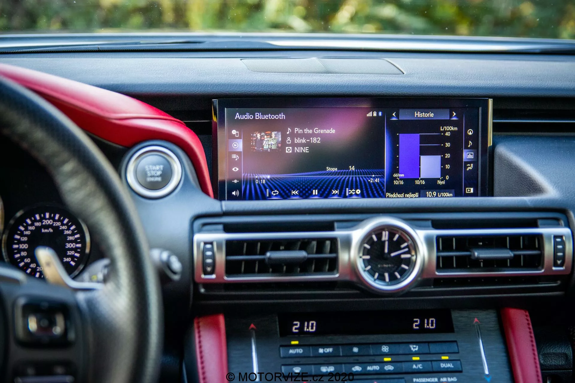 Vue rapprochée depuis le siège du conducteur d'une Lexus RC 2016, mettant en avant l'écran d'infodivertissement moderne avec des éléments tels que le nom de la piste, l'artiste et les niveaux audio, centré dans le cadre. Sous l'écran se trouvent les commandes de climatisation et une horloge analogique circulaire. Le tableau de bord présente un design de luxe ou sportif avec des matériaux contrastants, possiblement en cuir avec des coutures rouges sur un fond plus sombre. On aperçoit partiellement le compteur de vitesse et le compte-tours, ainsi que des commandes supplémentaires de la voiture en dessous des commandes de climatisation.