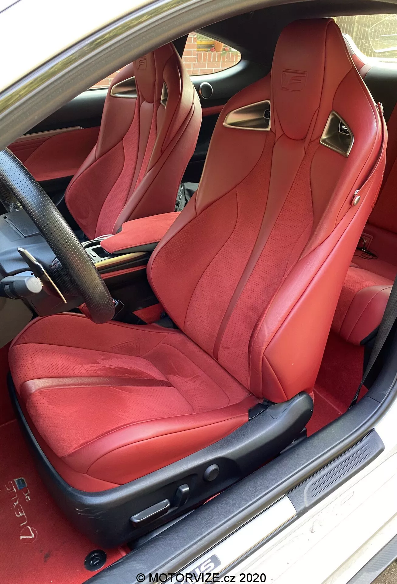 Vue de l'intérieur d'une Lexus RC 2016, montrant le luxueux siège passager avant en cuir rouge avec des renforts latéraux sportifs et un appui-tête intégré, vu du côté conducteur. Le siège présente des coutures complexes et des commandes réglables électriquement, complétées par des accents métalliques. La porte de la voiture est ouverte, révélant la plaque de seuil de porte marquée, avec un aperçu du pavé en briques résidentiel à l'extérieur.