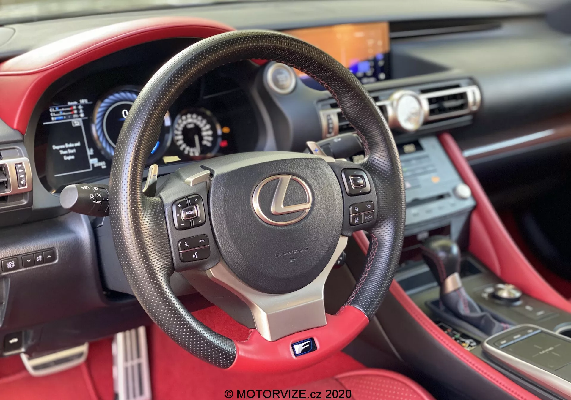 Vue en première personne de l'intérieur du Lexus RC 2016, se concentrant sur un volant à trois branches en cuir avec boutons de commande, palettes de changement de vitesse et badge argenté 'F' Sport. Tableau de bord numérique partiellement visible et console centrale avec écran d'info-divertissement et commandes de climatisation. Intérieur en cuir rouge saisissant ajoutant une sensation premium.