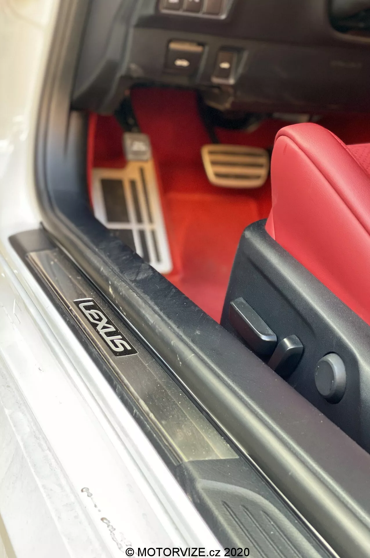 Vue rapprochée de l'intérieur d'une Lexus RC 2016, mettant en lumière le seuil de porte métallique avec plaque 'LOVIS', la zone de plancher recouverte de tapis rouge, les pédales en aluminium de style sportif, le panneau inférieur de la planche de bord, et le joint en caoutchouc noir de la porte. Photo prise par MOTORVIZE.cz 2020.