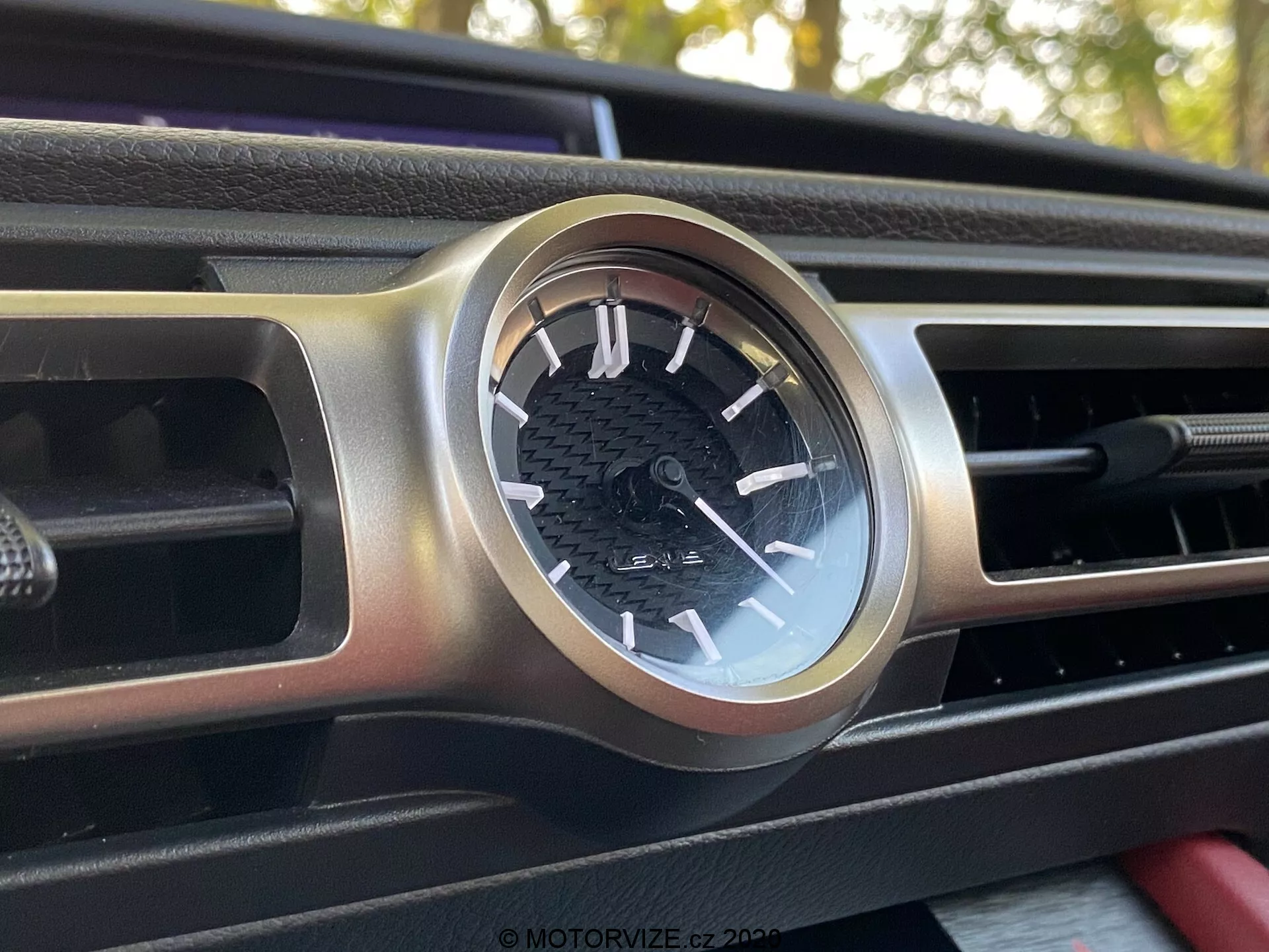 Vue rapprochée et légèrement inclinée d'une horloge analogique circulaire sur le tableau de bord de la Lexus RC 2016, enchâssée dans un boîtier en métal brossé. L'horloge présente un cadran noir avec des repères blancs, des chiffres des heures et une piste des minutes, positionnée au centre d'une grille de ventilation étendue horizontalement. Le tableau de bord affiche un motif noir texturé, possiblement en fibre de carbone. Les éléments d'arrière-plan sont flous, centrant l'attention sur le design détaillé de l'horloge.