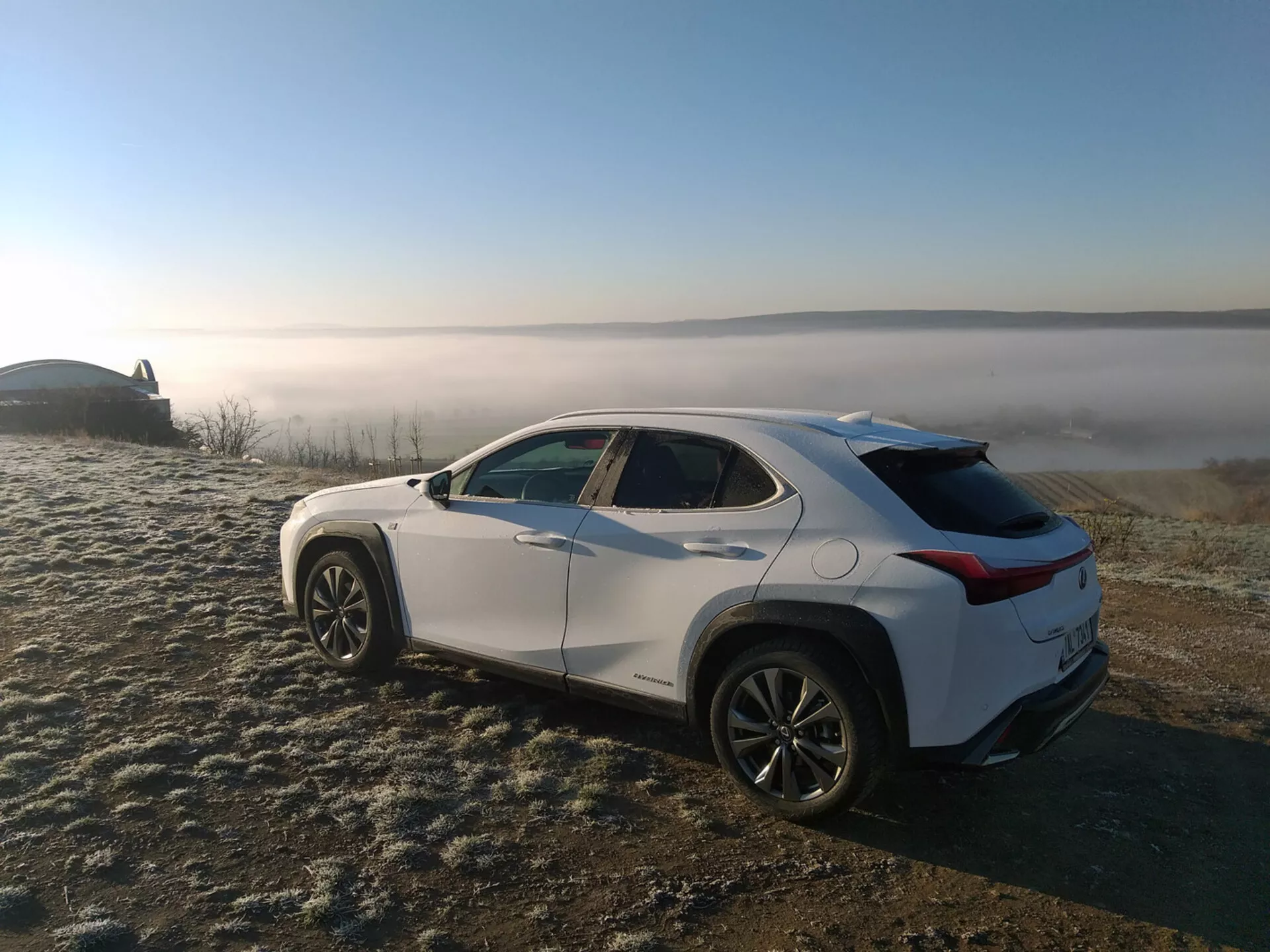 SUV crossover Lexus UX 2019 garé sur de l'herbe couverte de givre avec un arrière-plan de paysage brumeux, montrant le profil du véhicule à partir d'un angle arrière de trois quarts mettant en évidence les lignes de caractère latérales, les arches de roue, la ligne de toit effilée et les jantes en alliage.