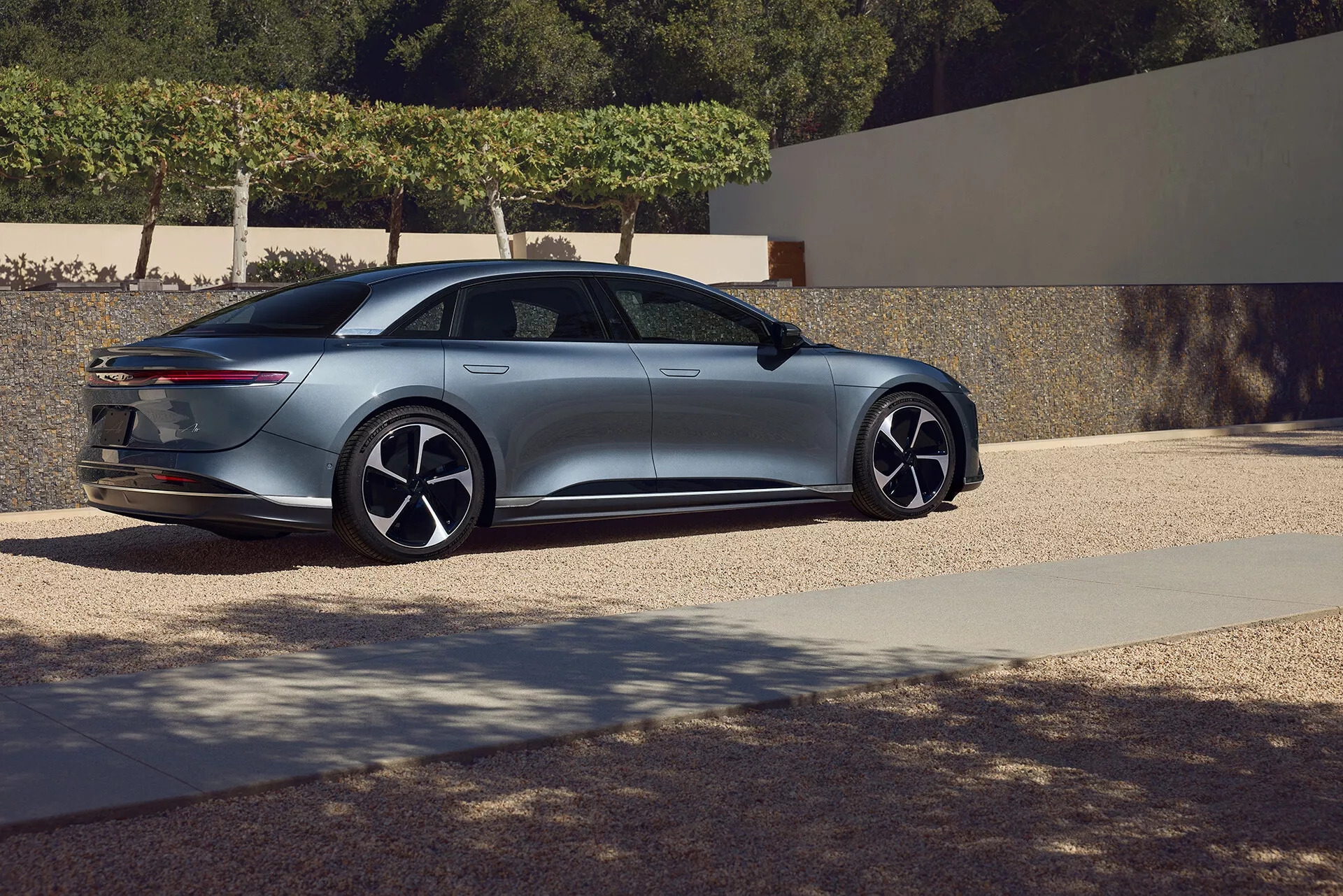 Vue latérale arrière trois quarts de la berline Lucid Air 2022 de première génération. La prise de vue en contre-plongée met en valeur le design fluide et aérodynamique du véhicule, avec une ligne de toit lisse se prolongeant dans un coffre de style fastback doté de feux arrière étroits et horizontaux. Le profil latéral présente de grandes roues en alliage à plusieurs rayons et des poignées de porte affleurantes. Les rétroviseurs latéraux fins et le capot incliné suggèrent des fonctionnalités technologiques avancées, renforçant l'impression de luxe et de modernité de la voiture. Le cadre semble être une zone résidentielle, accentuant l'esthétique premium.