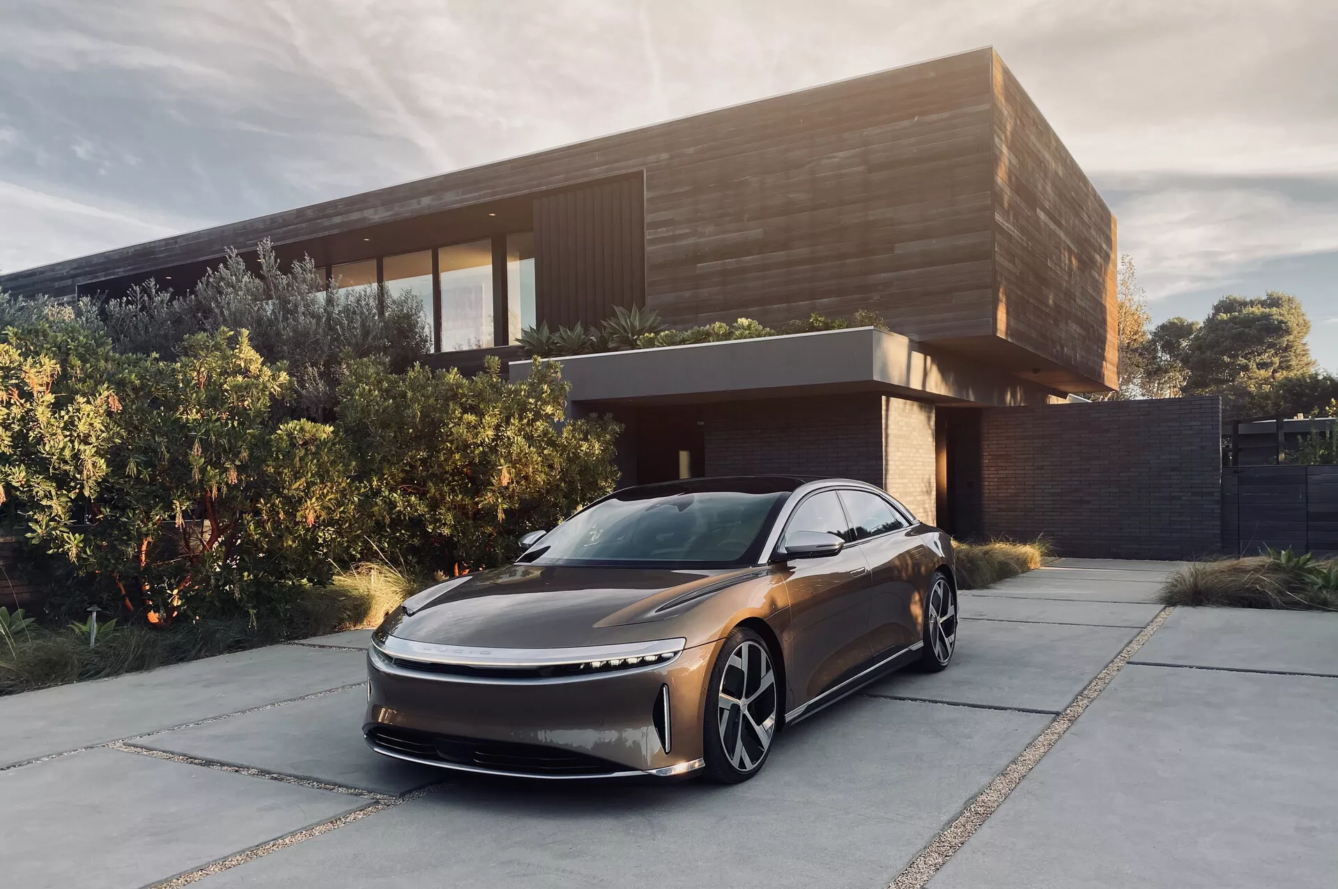 Image de la Lucid Air 2022, une berline moderne, garée devant une maison contemporaine. Vue sous un angle bas de trois quarts avant, montrant son design aérodynamique et élégant avec un capot allongé et une grille subtile. On distingue les phares à LED fins, les grandes roues distinctives et l'absence de rétroviseurs latéraux, ce qui suggère un système de vision arrière par caméra. La couleur foncée métallisée et l'environnement sophistiqué accentuent son allure luxueuse, laissant supposer qu'il s'agit d'un véhicule électrique destiné à une clientèle aisée.