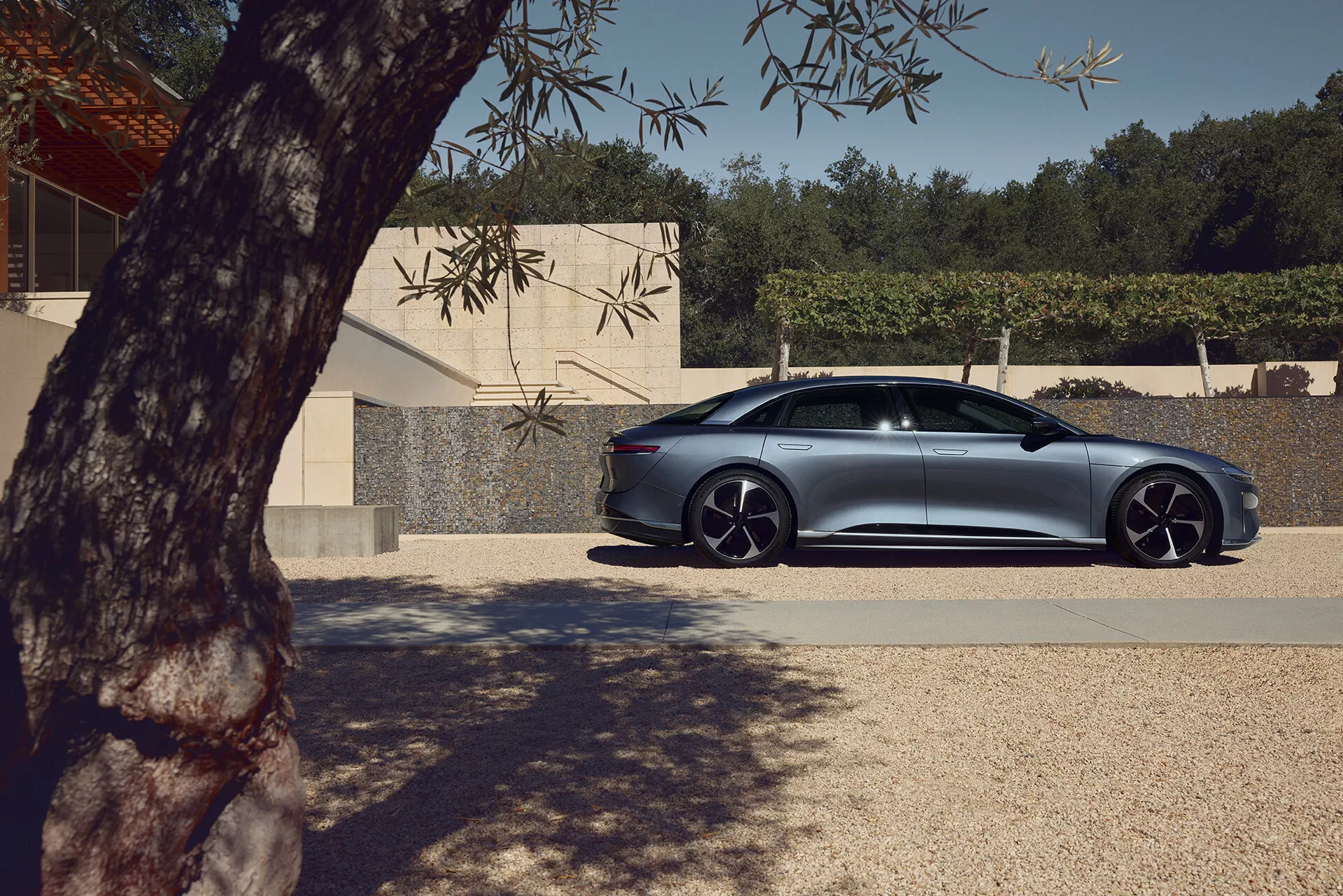 Vue de profil d'une berline à quatre portes Lucid Air 2022 de première génération stationnée à l'extérieur. Le côté passager de la berline luxueuse et performante est visible, mettant en évidence les lignes lisses de la carrosserie, le design des portes et des fenêtres, et les roues en alliage distinctives avec des pneus à profil bas. Partiellement ombragé par le feuillage d'un arbre, avec une lumière naturelle soulignant la finition métallique. Un bâtiment moderne à l'arrière-plan met en valeur le design élégant et aérodynamique de la voiture avec des caractéristiques avancées telles que les poignées de porte affleurantes et les rétroviseurs aérodynamiques pouvant indiquer des clignotants intégrés ou une détection d'angle mort.