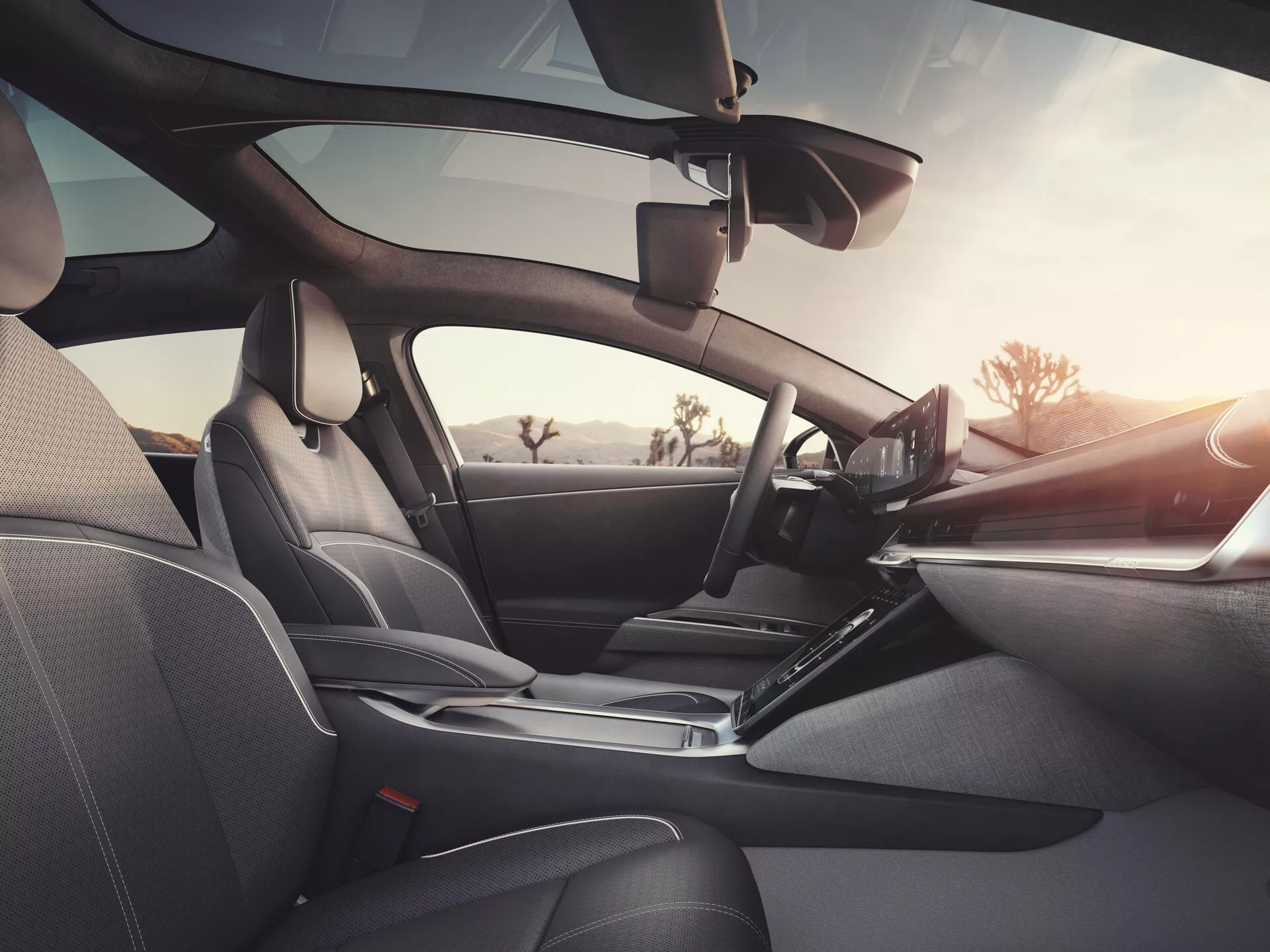 Vue intérieure de la Lucid Air 2022 (1ère génération) depuis la perspective du passager arrière, montrant des sièges en cuir foncé avec des coutures détaillées, un tableau de bord élégant avec un volant à gauche comprenant des commandes intégrées, un affichage d'infodivertissement tactile proéminent, un levier de changement de vitesse ergonomique, des panneaux de porte avec commandes électroniques et haut-parleurs, rétroviseur visible et possible dispositif d'assistance au conducteur près du pare-brise, mettant en avant un design luxueux, une technologie avancée et un confort accru.