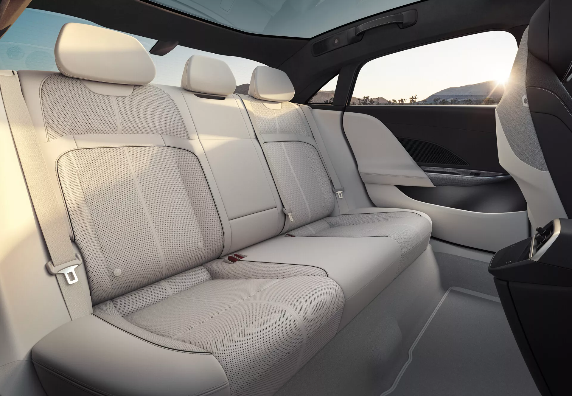 Vue intérieure de la Lucid Air 2022, se concentrant sur les sièges passagers arrière. Les sièges sont rembourrés en cuir de couleur claire, offrant une sensation d'aération et d'espace. La banquette arrière peut accueillir trois passagers avec des appuie-tête assortis et des ceintures de sécurité visibles. Les panneaux de porte se mêlent parfaitement au matériau et au design des sièges, comportant des grilles de haut-parleurs intégrées. Une ligne de fenêtre haute et un grand toit panoramique teinté rehaussent le sentiment d'intimité et de protection. La garniture intérieure montre un mélange de rembourrage clair et d'éléments plus sombres, probablement de matériaux de haute qualité.
