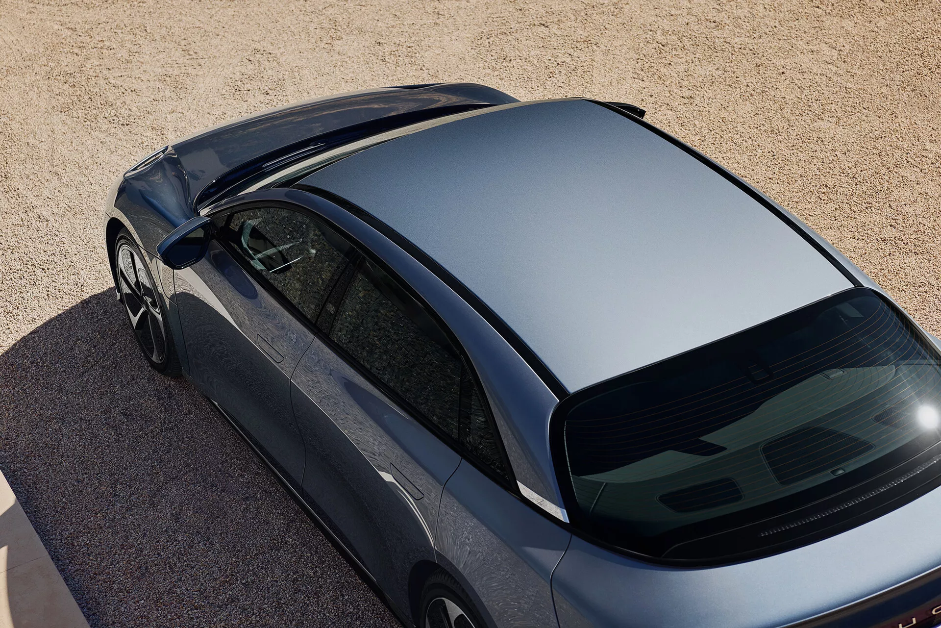 Vue aérienne oblique d'une voiture Lucid Air 2022 de première génération, de couleur métallique foncée, stationnée sur une surface de petits cailloux ou de sable grossier. La prise de vue capture la vue vers le bas, en se concentrant sur le toit profilé, la lunette arrière inclinée et une partie du coffre. Les caractéristiques comprennent des fenêtres sans cadre, des rétroviseurs latéraux profilés et l'absence d'accessoires de toit et d'essuie-glace arrière, suggérant un design aérodynamique et moderne.