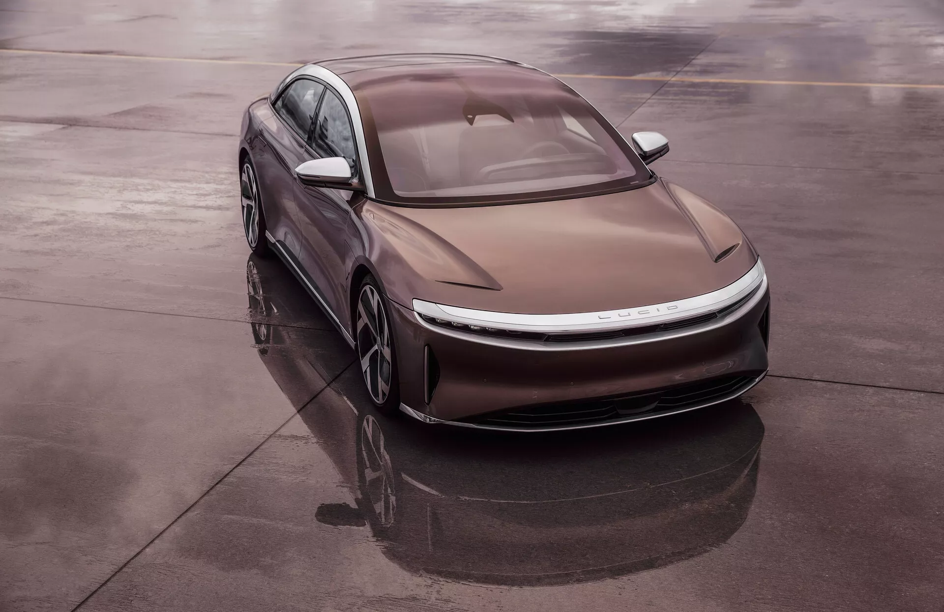 Photo en plongée d'une berline moderne Lucid Air 2022 de 1ère génération, positionnée en diagonale sur une surface réfléchissante comme l'asphalte mouillé, offrant une vue complète du quart avant gauche du véhicule. Le design aérodynamique élégant présente des courbes douces, des phares LED fins et un capot profilé avec des lignes marquées. La ligne de toit de type fastback et le verre affleurant améliorent l'efficacité aérodynamique. La nature électrique du véhicule est suggérée par l'absence de grille traditionnelle, un système d’admission d'air intégré, et des roues aérodynamiques légères.
