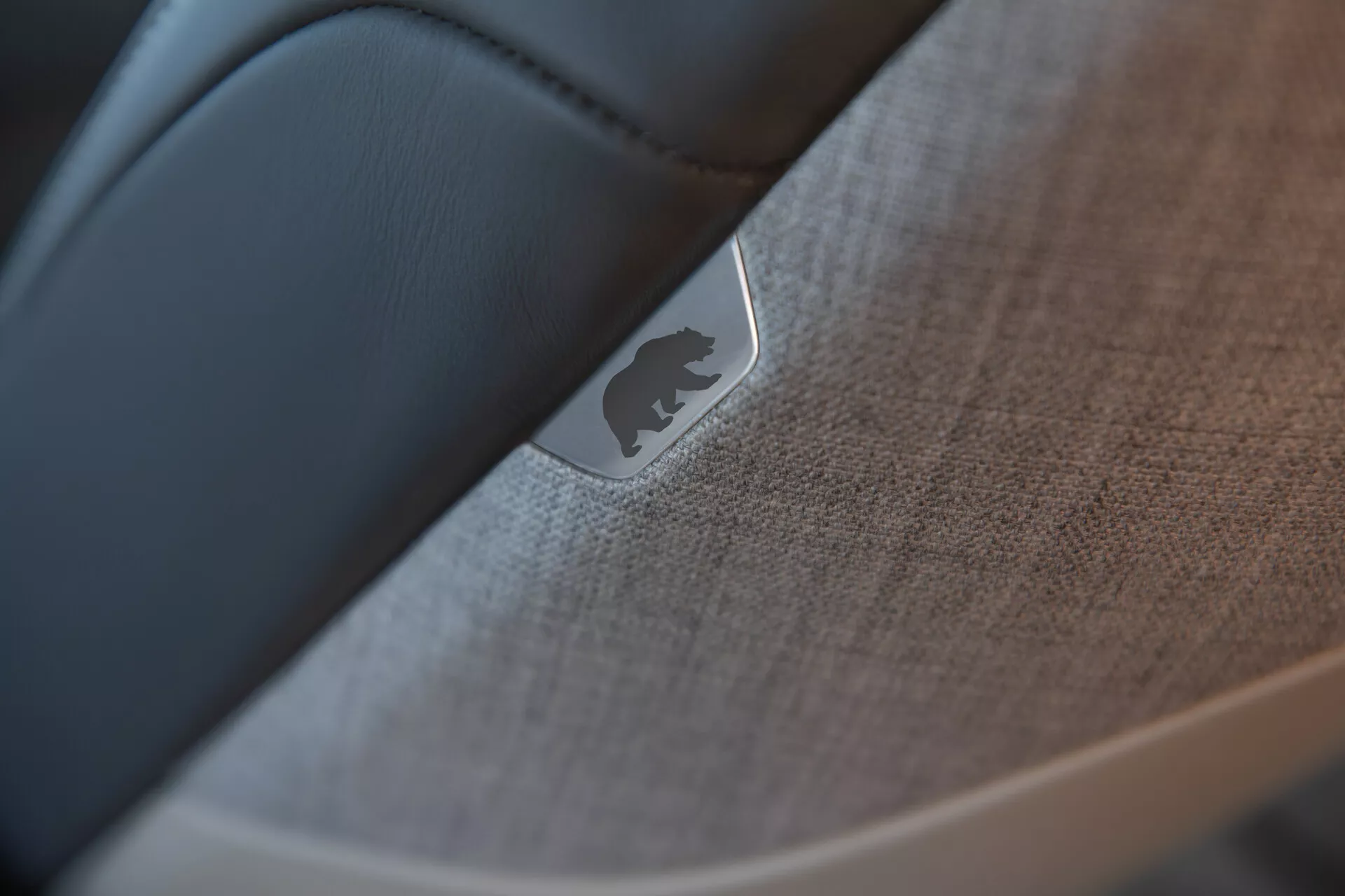 Gros plan, image à mise au point sélective de l'intérieur du véhicule Lucid Air 2022 (1ère génération) mettant en avant le détail d'une porte ou du tableau de bord. Un emblème ou badge métallique réfléchissant est bien visible, placé sur un tissu texturé ou un plastique doux au toucher avec des détails de coutures. Un matériau lisse et sombre, probablement du cuir noir, contraste avec les éléments texturés, montrant l'artisanat et la haute qualité de la conception intérieure de la voiture.