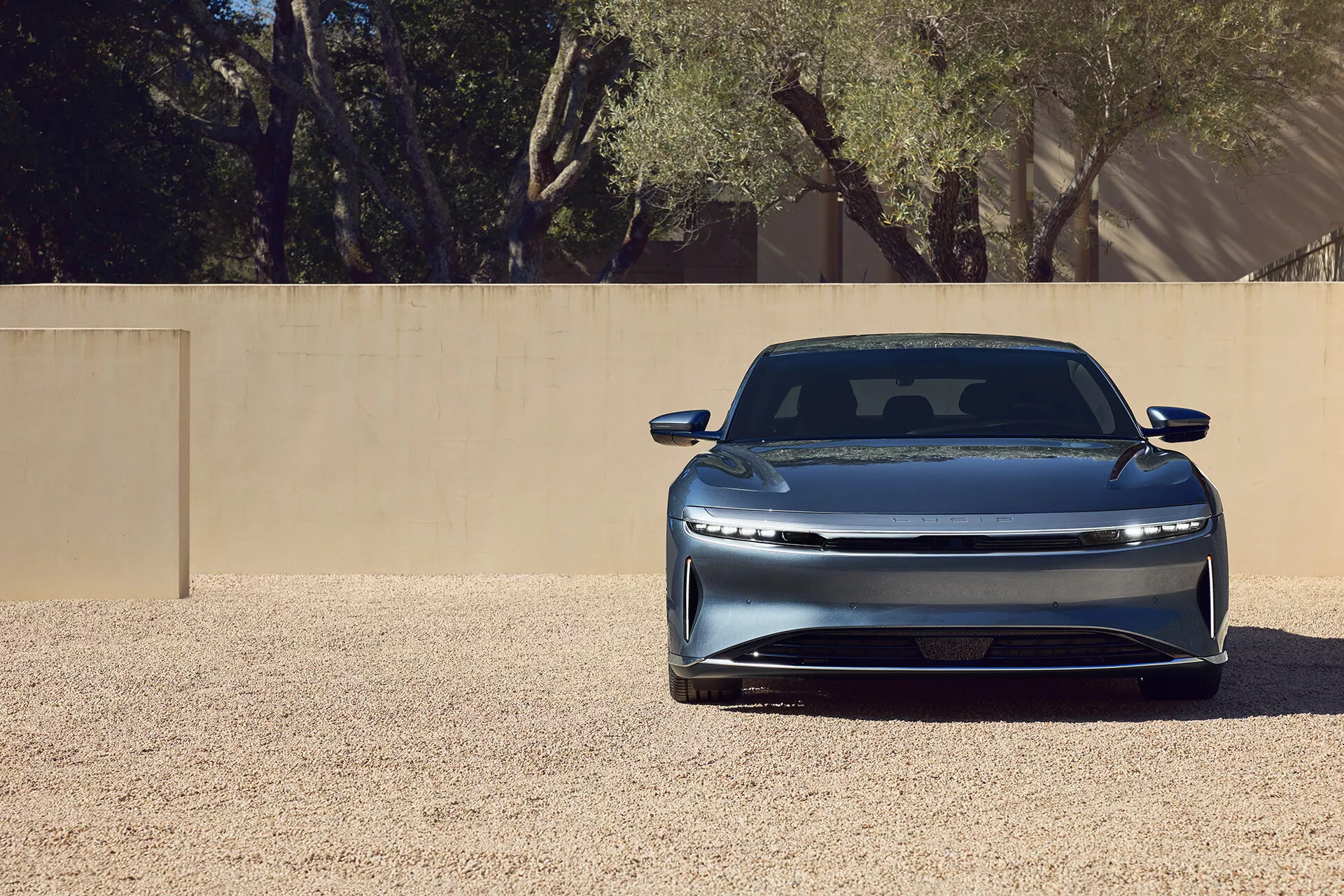 Gros plan de l'avant de la Lucid Air 2022 de première génération, mettant en avant sa grille fermée élégante, ses phares LED horizontaux, son pare-chocs avant intégré, son capot sculpté avec une ligne de contour et sa finition de peinture métallique. La voiture est positionnée au centre sur une surface de gravier sous un éclairage brillant, suggérant une aérodynamisme avancé et un design moderne de luxe.