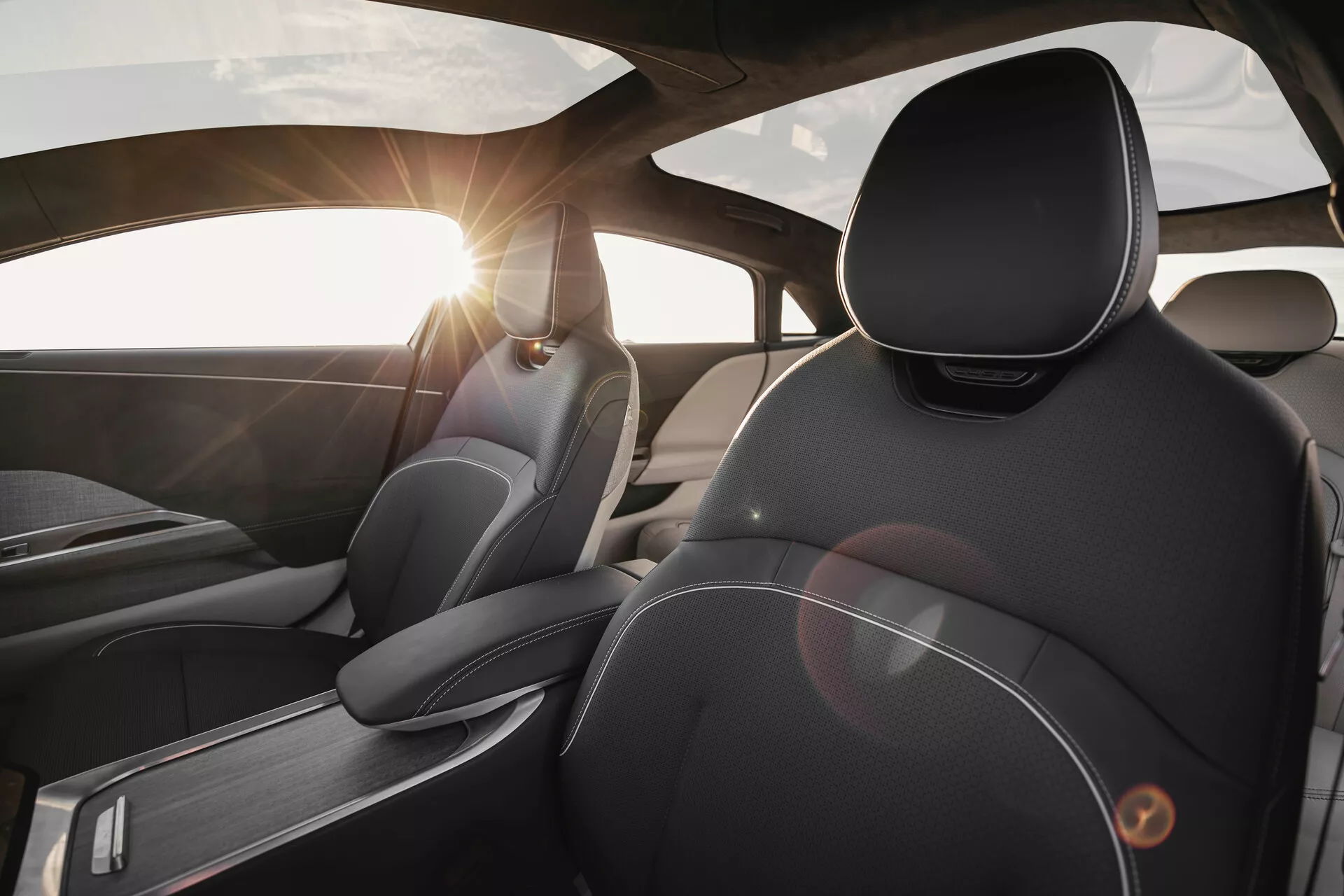 Vue intérieure de la Lucid Air 2022 depuis une perspective du siège arrière, centrée sur le siège du conducteur. L'accent est mis sur les matériaux et le design premium, avec des coutures détaillées sur le cuir, des appuis-tête ergonomiques, des accents en métal brossé et un effet de lens flare du soleil à travers le pare-brise.