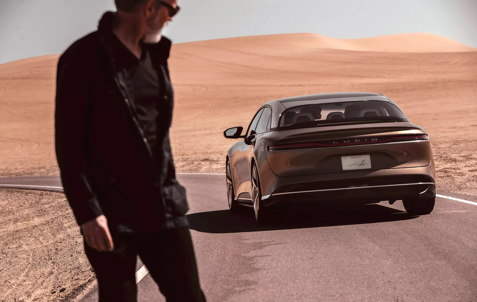 Une berline élégante et moderne Lucid Air 2022 (1ère Génération) garée dans un paysage désertique, capturée d'un angle bas en se concentrant sur les trois quarts arrière. La couleur de carrosserie bronze métallique et les feux arrière horizontaux proéminents soulignent le design contemporain de la voiture. Une bande chromée réfléchissante court sur la largeur du coffre sous les feux. Le badge du fabricant est centré sur le coffre. Le pare-chocs arrière intégré est muni de capteurs de stationnement, ajoutant à l'apparence luxueuse. Une silhouette floue se tient au premier plan, suggérant un mouvement ou une activité près du véhicule.