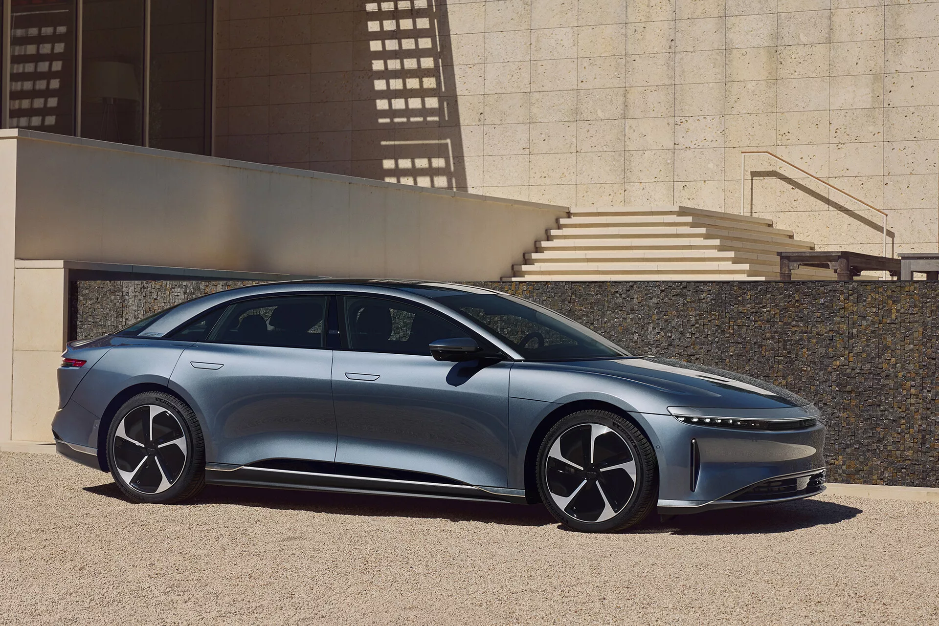 Une vue de profil latéral de la Lucid Air 2022, une berline moderne et élégante, capturée en extérieur dans un cadre urbain avec des éléments architecturaux projetant des ombres. Le côté gauche de la voiture montre son design aérodynamique avec un toit bas, des poignées de portes affleurantes, de grandes roues à plusieurs rayons et des rétroviseurs latéraux fins. L'image met en valeur la longueur de la voiture, du pare-chocs avant au pare-chocs arrière, avec un pilier A incliné vers l'avant donnant une impression d'intérieur spacieux et une ligne de caractère dynamique sur la carrosserie.