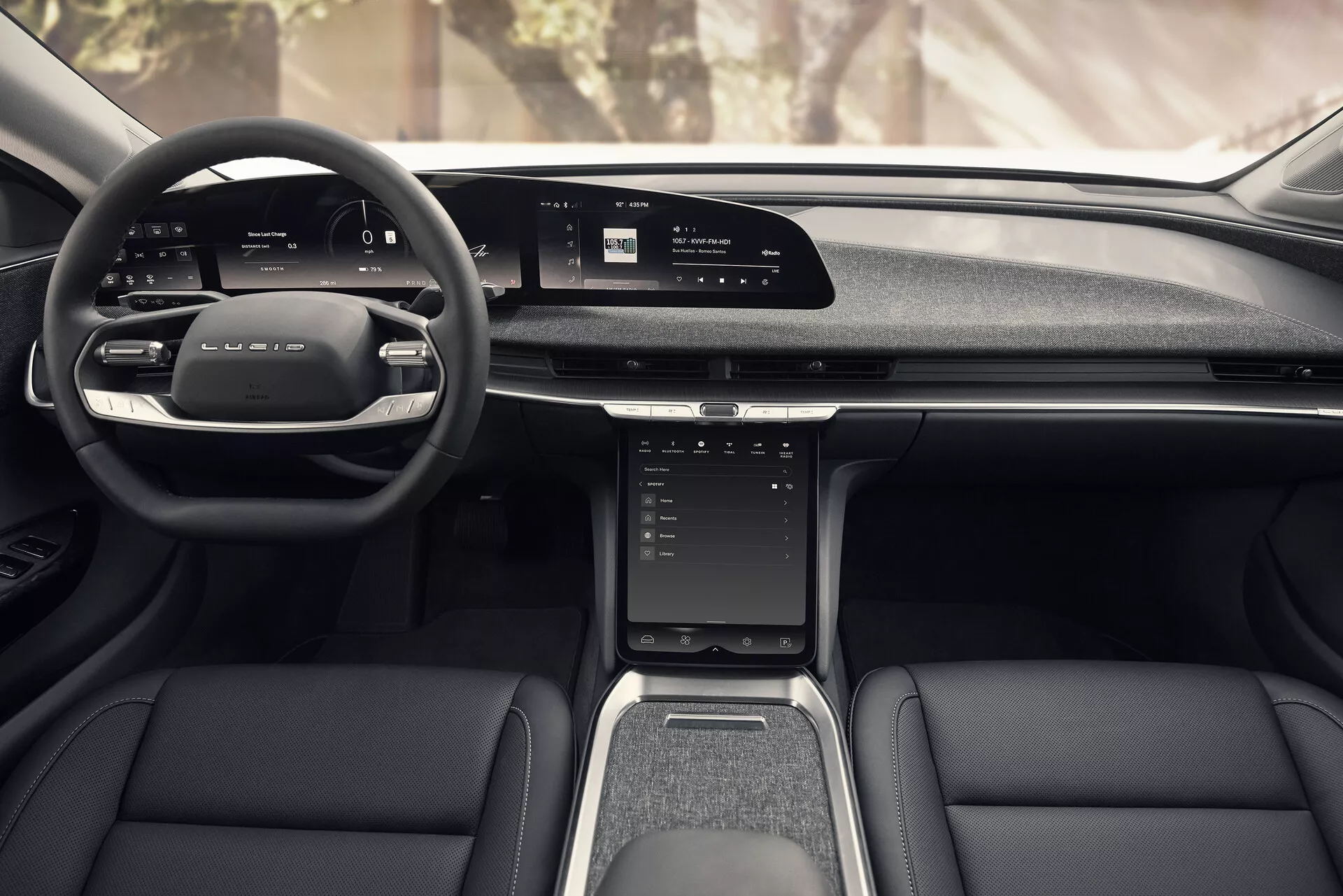 Vue depuis le siège central arrière de l'intérieur du Lucid Air 2022, présentant un design moderne avec volant multifonction en cuir, large écran digital sur le tableau de bord, écran tactile vertical sur la console centrale, palettes de sélection manuelle des vitesses, sièges avant élégants en cuir et tissu, tunnel central avec levier de vitesses et porte-gobelets, et panneaux de portes avec poches de rangement et grilles de haut-parleurs.