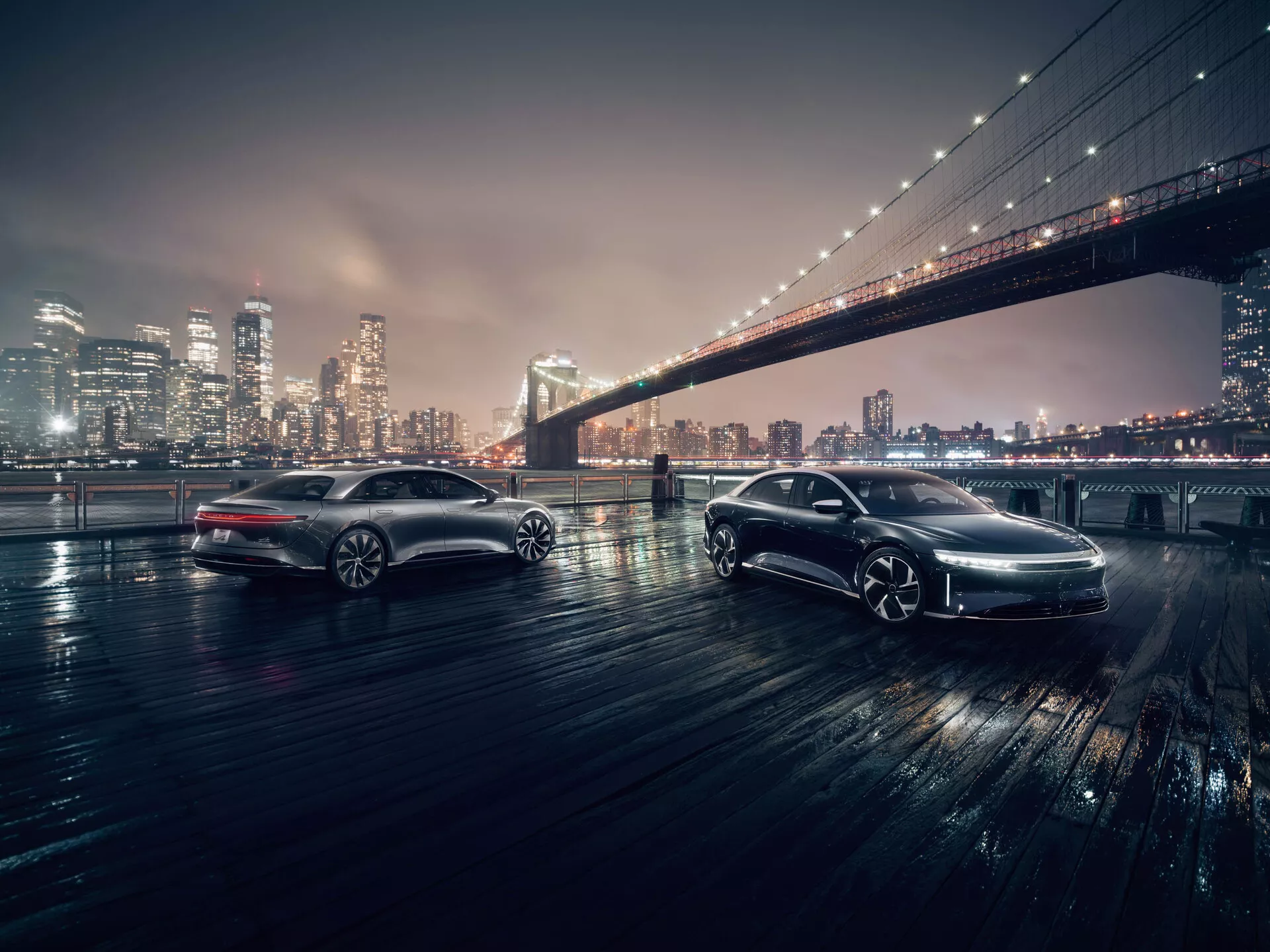 Deux voitures élégantes dans un cadre urbain nocturne avec le pont de Brooklyn emblématique en arrière-plan. Les voitures sont photographiées sous un angle bas, mettant en évidence les détails de leur design. La voiture au premier plan, orientée vers la caméra, présente sa calandre avant, ses phares à LED et ses essuie-glaces à capteur de pluie. La voiture en arrière-plan, parallèle à la caméra, révèle son profil latéral, ses jantes en alliage et sa peinture de haute qualité. Les deux véhicules sont sur une surface mouillée qui reflète leur éclairage à LED, suggérant des technologies avancées telles que l'ABS et le contrôle de traction.