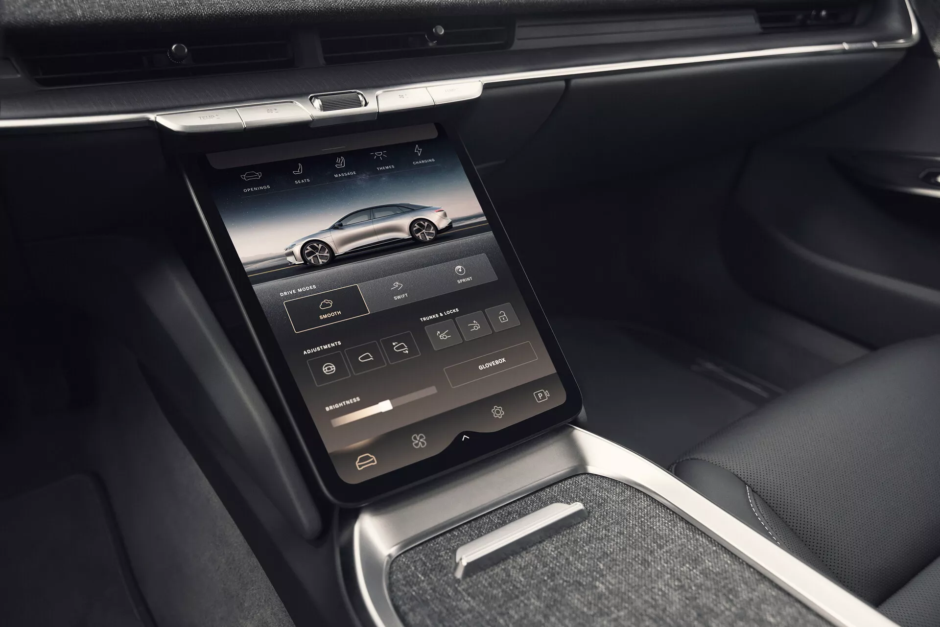 Vue intérieure de la Lucid Air 2022 (1ère génération) présentant la console centrale avec un écran tactile intégré monté verticalement, avec des icônes pour la navigation, le téléphone, les médias, la climatisation et les réglages du véhicule. La photo, prise d'une perspective légèrement inclinée vers la gauche, met en avant des fonctionnalités technologiques avancées avec des bouches d'aération au-dessus de l'écran, des garnitures brillantes en chrome ou en métal, un tableau de bord élégant et un mélange de finitions en cuir et en métal. L'image détaille également une petite étagère sous l'écran tactile, un levier de vitesse métallique et des accoudoirs et côtés de sièges en cuir, soulignant un intérieur luxueux et confortable.