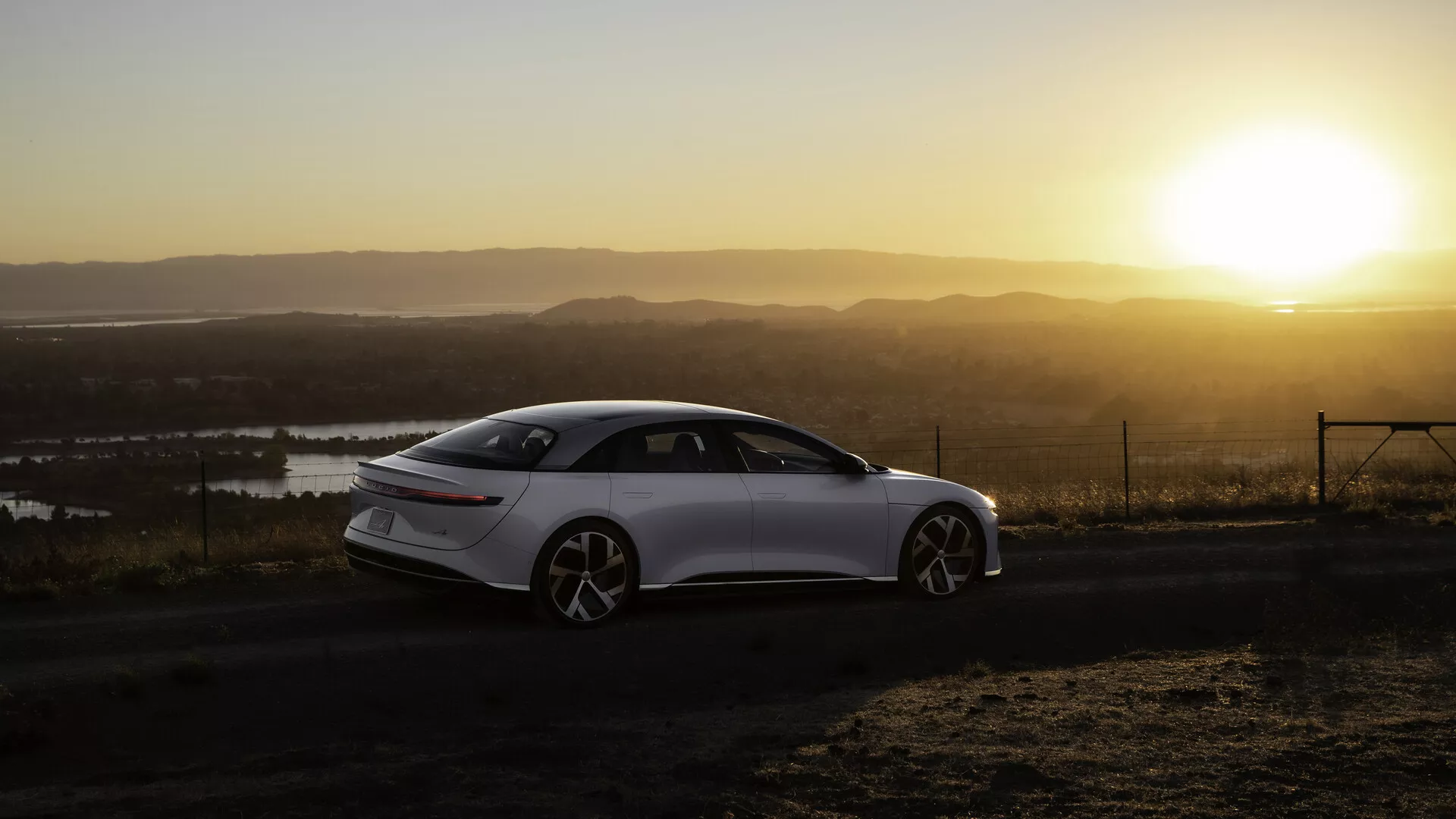 Lucid Air 2022 (1re Génération) photographié sous un angle arrière latéral au lever ou au coucher du soleil avec un soleil chaud en arrière-plan, positionné sur le côté gauche du cadre. La berline quatre portes élégante et moderne présente des lignes fluides, de grandes roues à plusieurs rayons et des poignées de porte rétractables. Le paysage comprend des plans d'eau et un point de vue élevé. Les feux arrière sont étroits et allongés horizontalement, avec la lumière du soleil créant un effet de lueur chaude et de flare sur la scène.