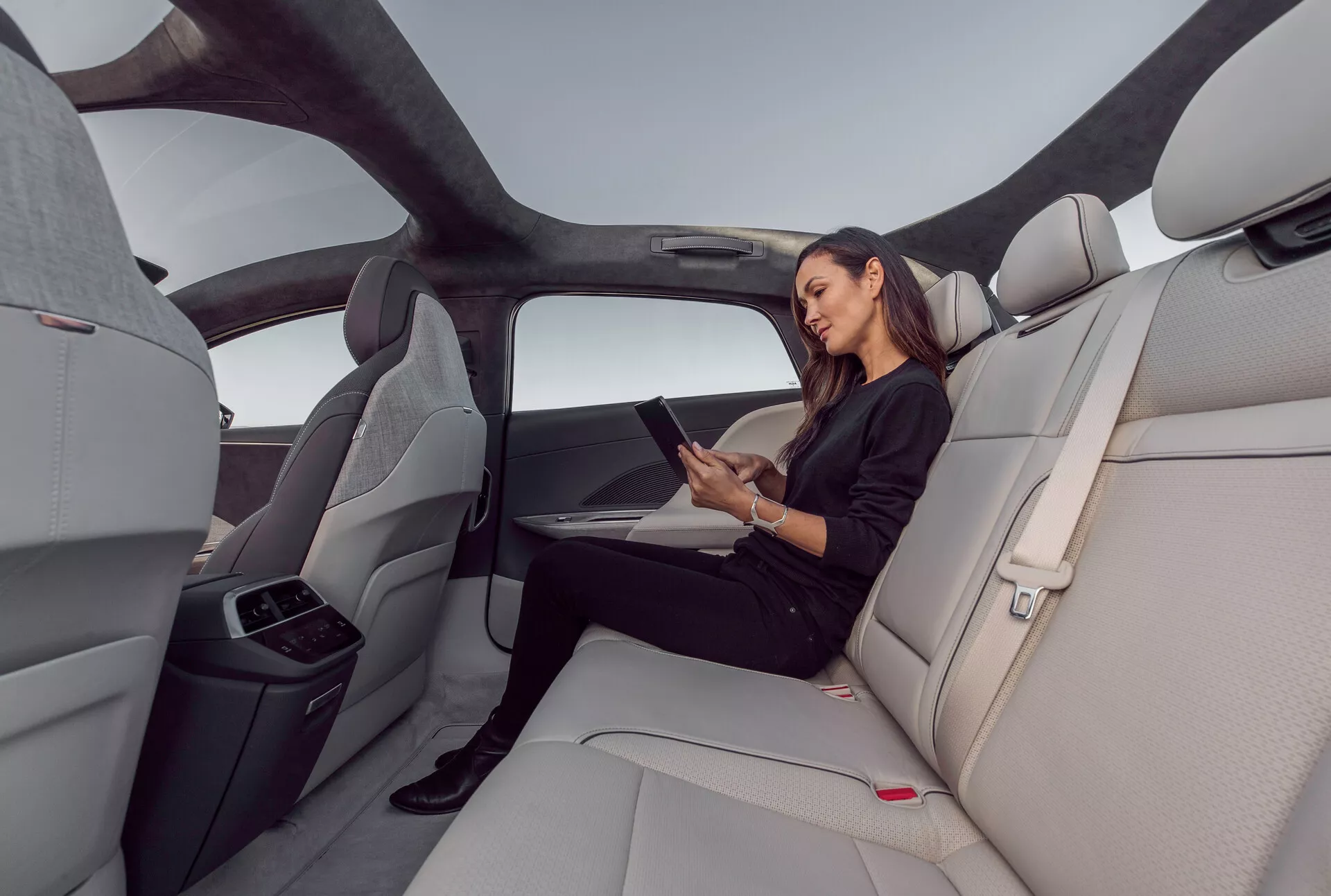 Vue intérieure de la zone des sièges arrière du Lucid Air 2022, mettant en avant la cabine spacieuse et luxueuse avec des sièges en cuir contournés gris clair. L'image capture une vue grand-angle du côté droit, montrant le toit panoramique s'étendant sur l'espace arrière, les designs ergonomiques des sièges et un accoudoir central replié avec porte-gobelets. Une femme est assise confortablement en interagissant avec une tablette, indiquant des fonctionnalités potentielles de connectivité. Intérieur élégant et minimaliste avec des boutons de libération de ceinture de sécurité rouges et des lignes horizontales sur le tableau de bord et les panneaux de porte, améliorant la sensation de largeur et d'ouverture.