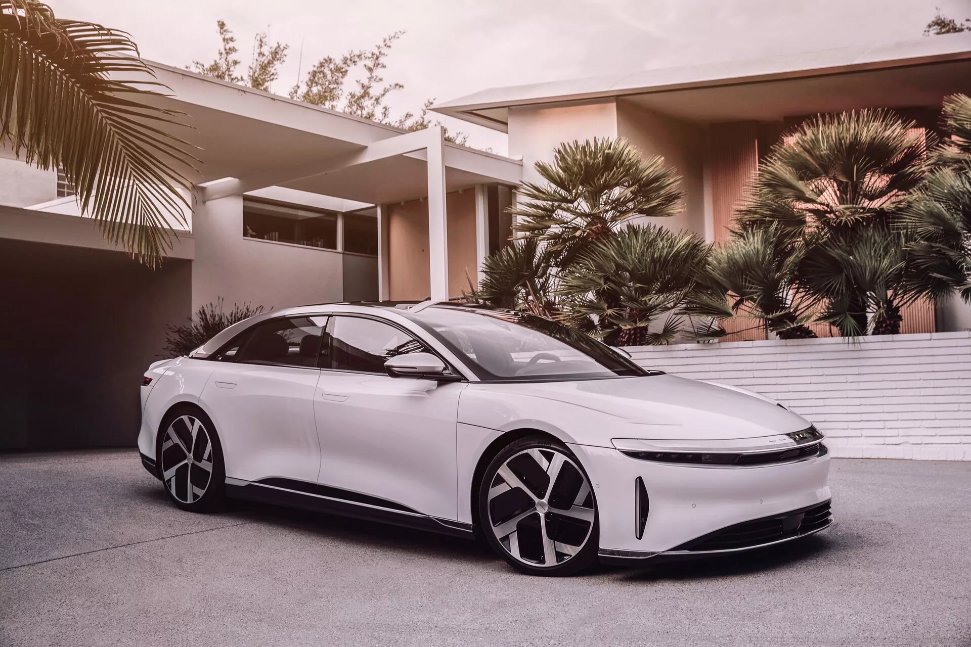 L'image montre une Lucid Air 2022 de première génération, une berline blanche moderne et élégante avec une position basse et large et une ligne de toit de type coupé. Capturée sous un angle bas, cette prise de vue accentue son design aérodynamique, ses grandes jantes distinctives, ses phares à LED et ses rétroviseurs latéraux profilés, suggérant des technologies avancées d'assistance à la conduite. La calandre minimaliste laisse supposer des technologies de propulsion électrique ou hybride. Le design futuriste des jantes comporte des caches aérodynamiques, et l'apparence du véhicule est rehaussée par des vitres teintées, des portes sans cadre et des poignées de porte affleurantes élégantes. Une vue partielle du toit panoramique suggère un habitacle luxueux et lumineux. L'arrière-plan montre une zone résidentielle de haut standing avec des palmiers, indiquant que la voiture est destinée à une clientèle aisée.