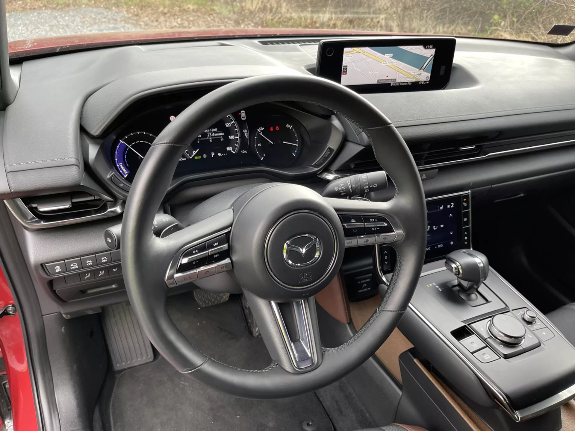 Vue intérieure de la Mazda MX-30 2020 du point de vue du conducteur, mettant en lumière des détails tels que le volant à trois branches enveloppé de cuir avec boutons de commande et palettes de changement de vitesse, tableau de bord numérique avec accents bleus, système d'infodivertissement avec écran tactile et navigation sur la console centrale, levier de vitesse sportif, bouton sélecteur de mode de conduite, bouton de contrôle supplémentaire pour le système d'infodivertissement, mélange luxueux de matériaux avec finitions noires et rouges, porte-gobelet et panneaux de contrôle sur la porte pour les rétroviseurs latéraux et les fenêtres, soulignant la technologie moderne et la conception ergonomique.