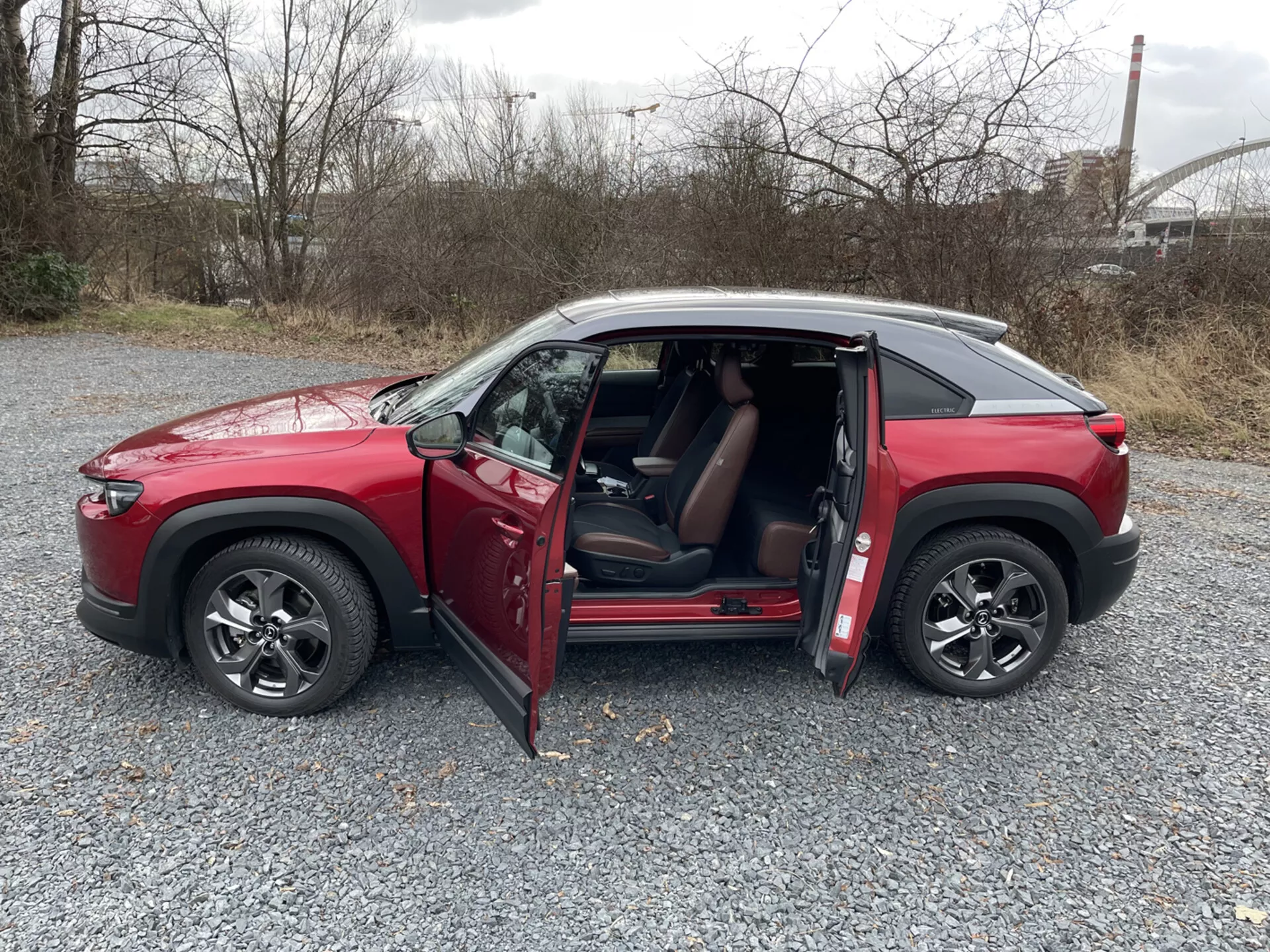 SUV Mazda MX-30 rouge quatre portes de 2020 en profil latéral sur gravier avec portes ouvertes, jantes en alliage à multiples branches, vitres teintées et toit de couleur contrastée sur un fond industriel et de verdure.