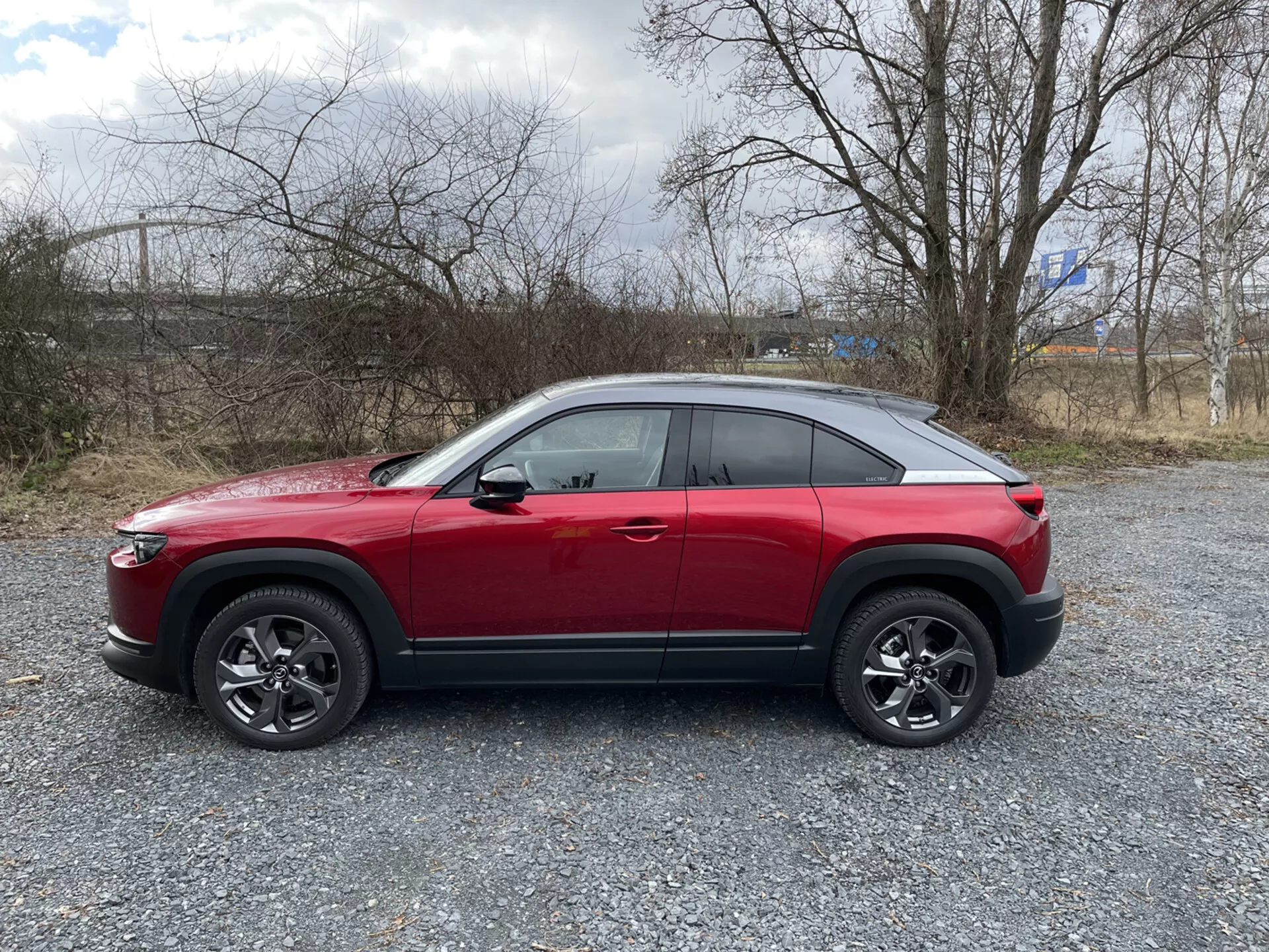 Profil latéral du SUV crossover rouge Mazda MX-30 2020 sur gravier avec végétation clairsemée et ciel nuageux, présentant une ligne de ceinture haute, une ligne de toit inclinée tendance coupé, une ligne de caractère dynamique, des roues en alliage à multiples branches avec garniture noire, et de possibles fonctionnalités technologiques avancées telles que l'ADAS, l'éclairage LED, l'entrée sans clé, le TPMS et le système d’infodivertissement.