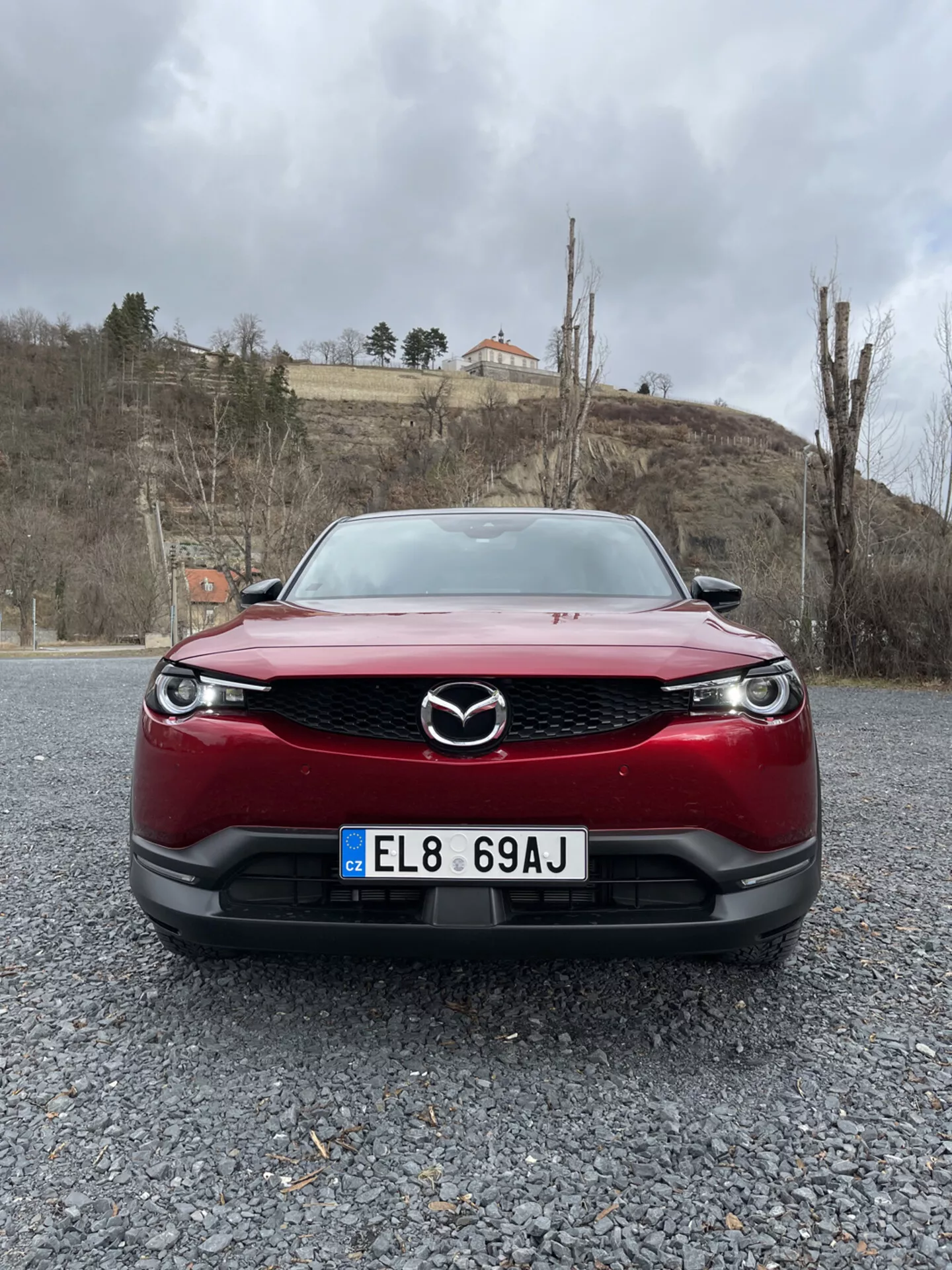 Vue de face d'une Mazda MX-30 de 2020 en rouge, mettant en avant la grille, le pare-choc et les phares LED de la voiture sur une surface de gravier contre un fond de ciel nuageux avec de la verdure et un bâtiment