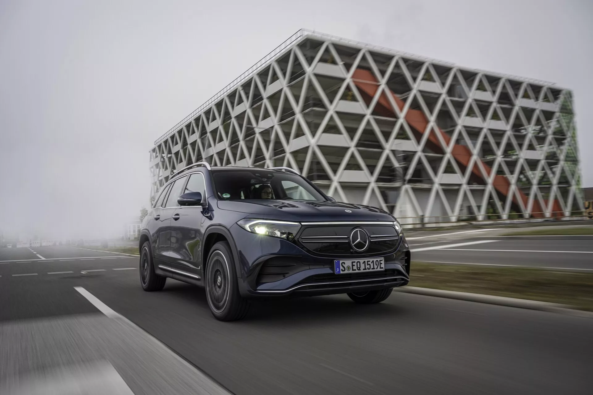 L'image montre un SUV Mercedes EQB 2021 à quatre portes en mouvement, capturé sous un angle bas et de côté. Le véhicule est positionné à gauche, affichant sa façade avant, son capot et son profil latéral jusqu'à la porte arrière. La roue avant de la voiture est légèrement tournée vers le spectateur, indiquant une manœuvre dynamique. Les caractéristiques visibles incluent la grille avant avec un emblème proéminent, des phares à LED, des feux de jour et un design aérodynamique. La finition métallisée de la peinture reflète la lumière, rehaussant sa surface polie. Les jantes en alliage ajoutent à l'esthétique, suggérant des caractéristiques de performance. L'arrière-plan comprend un bâtiment futuriste avec une façade géométrique, mettant en avant un environnement urbain et technologique qui correspond aux caractéristiques technologiques avancées du véhicule.