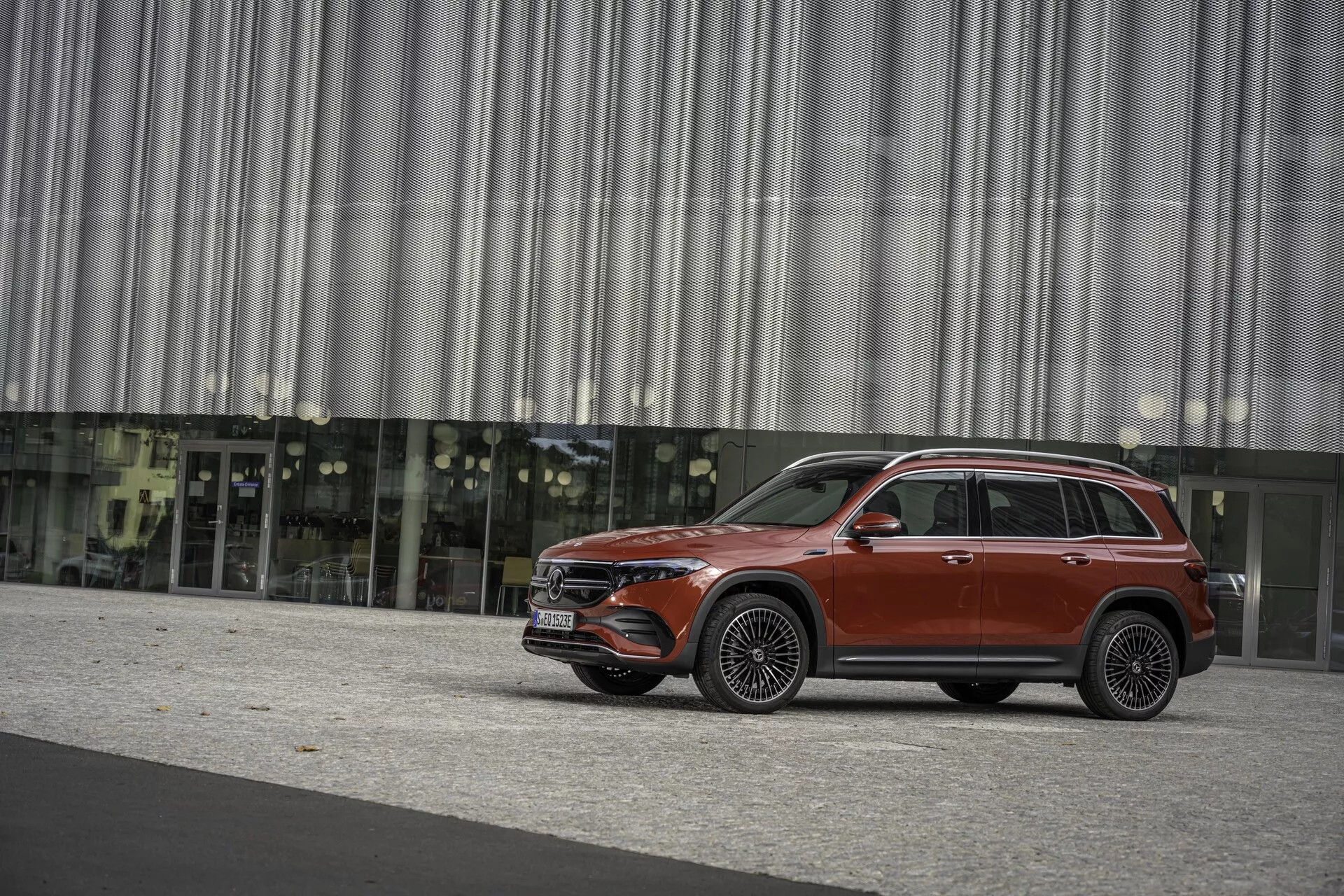 Profil latéral d'un SUV rouge, probablement le Mercedes EQB 2021, capturé sous un angle latéral, montrant toute sa longueur et ses éléments de design tels que les portes latérales, les fenêtres, les rétroviseurs, les roues et la carrosserie. Le véhicule se tient sur une surface pavée avec en arrière-plan un immeuble moderne avec une façade en verre réfléchissant et un mur-rideau métallique, suggérant un environnement urbain. L'image est prise sous la lumière naturelle avec des ombres douces, indiquant un ciel couvert ou une protection contre la lumière directe du soleil. Le SUV présente un design élégant, possiblement équipé de systèmes de navigation avancés, ADAS, systèmes d'infodivertissement, efficacité énergétique ou propulsion alternative et éléments de luxe comme un toit panoramique et des sièges en cuir.