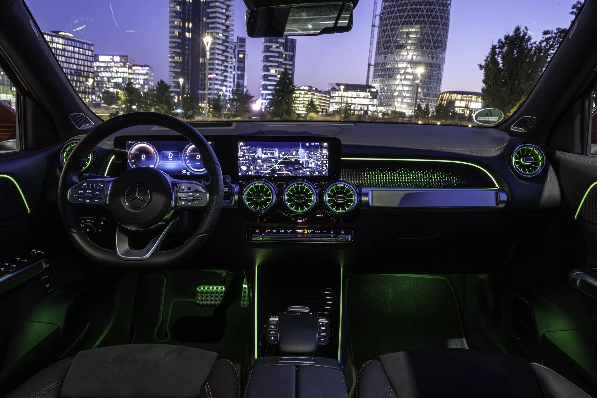 Vue intérieure de la Mercedes EQB 2021 depuis le siège arrière, montrant la cabine avant. Le volant ergonomique avec plusieurs boutons de contrôle, le fond plat et le tableau de bord numérique illuminé créent une ambiance high-tech. L'écran central d'infodivertissement et les aérateurs ronds illuminés ajoutent à l'attrait moderne. La console inférieure comporte un levier de vitesses moderne, entouré de commandes rétroéclairées en vert. Les sièges en cuir et l'éclairage ambiant vert créent une atmosphère futuriste. La vue extérieure de la ville visible à travers le pare-brise suggère un cadre urbain nocturne.