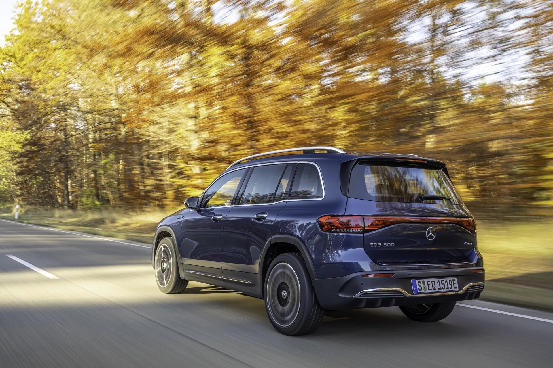 Vue dynamique d'une Mercedes EQB 2021 en mouvement sur une route bordée d'arbres. L'angle de trois quarts arrière montre le côté droit du SUV électrique avec un design moderne et élégant, finition métallisée et posture surélevée. Les points forts incluent les feux arrière, le pare-chocs arrière, les roues en alliage, les portières latérales et la ligne de toit inclinée. Le flou de mouvement suggère la vitesse, et le cadre lumineux de jour ajoute à l'esthétique haut de gamme.