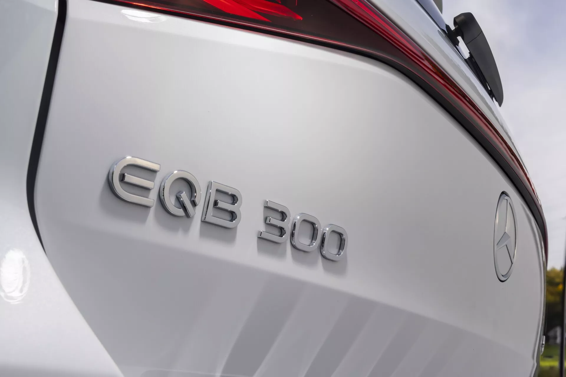 Gros plan en angle diagonal de l'arrière de la Mercedes EQB 300 2021, mettant en valeur le badge et l'emblème. L'image montre l'étoile à trois branches dans un cercle, la désignation du modèle 'EQB 300', une partie du hayon et le feu arrière gauche. L'angle de la caméra est en contre-plongée, mettant en avant la marque, les éléments de design moderne, la technologie LED et la qualité de construction premium de ce véhicule entièrement électrique. La carrosserie de la voiture est en finition blanche brillante, soulignant son attrait de luxe.