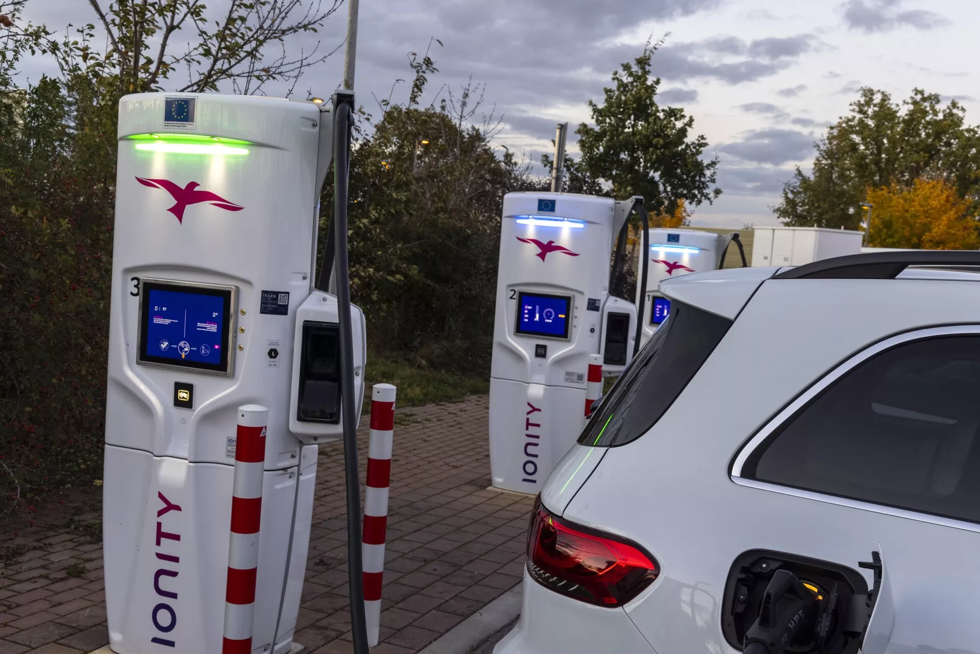 L'image représente une voiture blanche, partiellement visible depuis l'arrière gauche, en session de recharge électrique à une station de recharge. La prise de vue, réalisée sous un léger angle vers le côté conducteur, met en évidence le port de recharge ouvert situé au-dessus de la roue arrière et derrière la porte passager. Le feu arrière gauche, la vitre arrière et une partie du toit suggèrent une carrosserie de type berline ou SUV. En arrière-plan, deux bornes de recharge IONITY avec des interfaces à écran bleu et une lumière verte sur l'une d'elles indiquent une activité de recharge. L'éclairage ambiant suggère des conditions de soirée ou de ciel couvert, mettant en avant la voiture comme sujet principal dans une zone de recharge désignée.