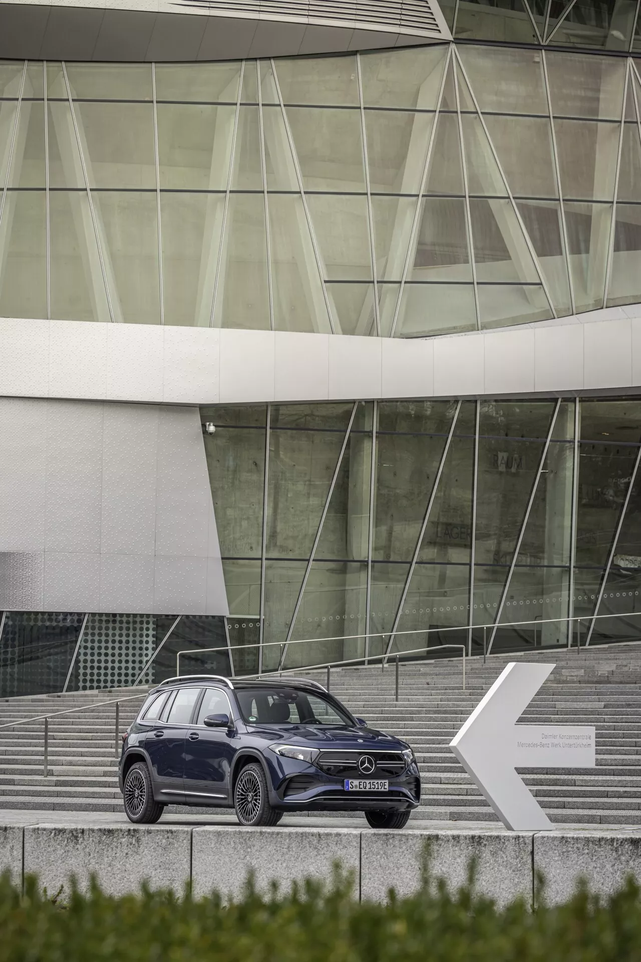 Profil latéral complet du SUV élégant Mercedes EQB 2021 positionné sur des marches menant à un bâtiment contemporain avec une façade en verre. Le SUV est centré et légèrement incliné vers la gauche, montrant son côté droit avec des lignes de design, des jantes en alliage à rayons multiples, des rétroviseurs latéraux, des fenêtres et des poignées de porte. La teinte foncée contraste magnifiquement avec les garnitures métalliques.