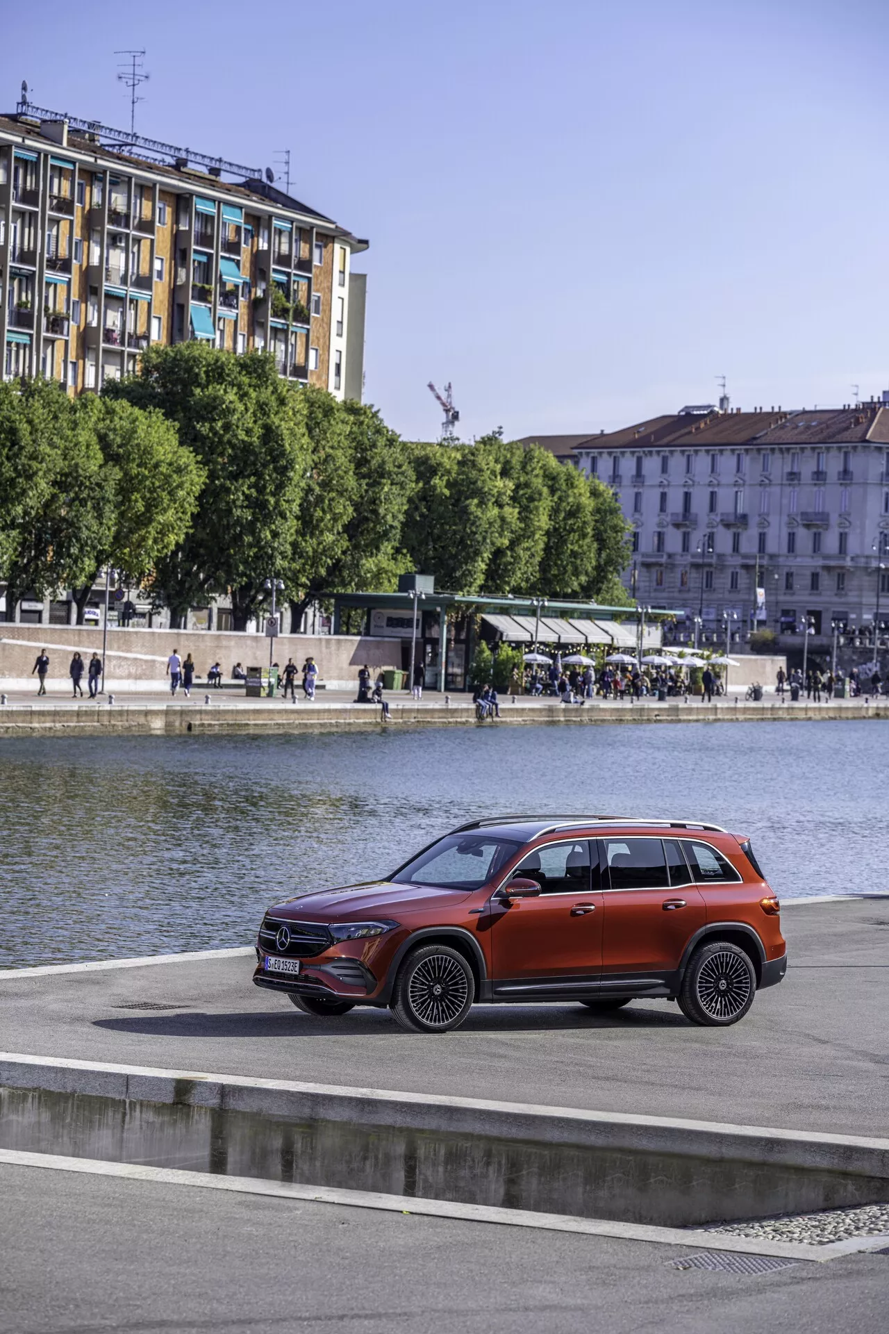 Vue latérale d'un SUV Mercedes EQB 2021 garé sur une promenade au bord de l'eau avec des bâtiments urbains et un arrêt de tramway en arrière-plan, mettant en valeur son design moderne, ses phares élégants, sa calandre avant proéminente, ses passages de roue bien définis et sa finition métallique brillante, vue d'un angle élevé.