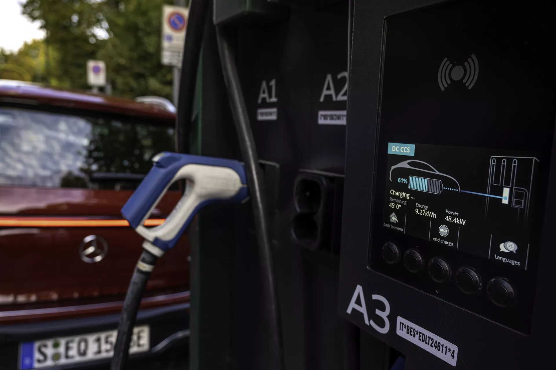 Vue rapprochée d'une station de recharge pour véhicules électriques avec une voiture en cours de chargement en arrière-plan. La station de recharge affiche des informations détaillées sur son écran numérique, telles que le statut de charge, l'énergie en kWh et la durée de la charge. La voiture, une berline compacte ou de taille moyenne rouge ou marron, est partiellement visible avec son feu arrière et le hayon, dans un cadre extérieur avec lumière naturelle et feuillage.