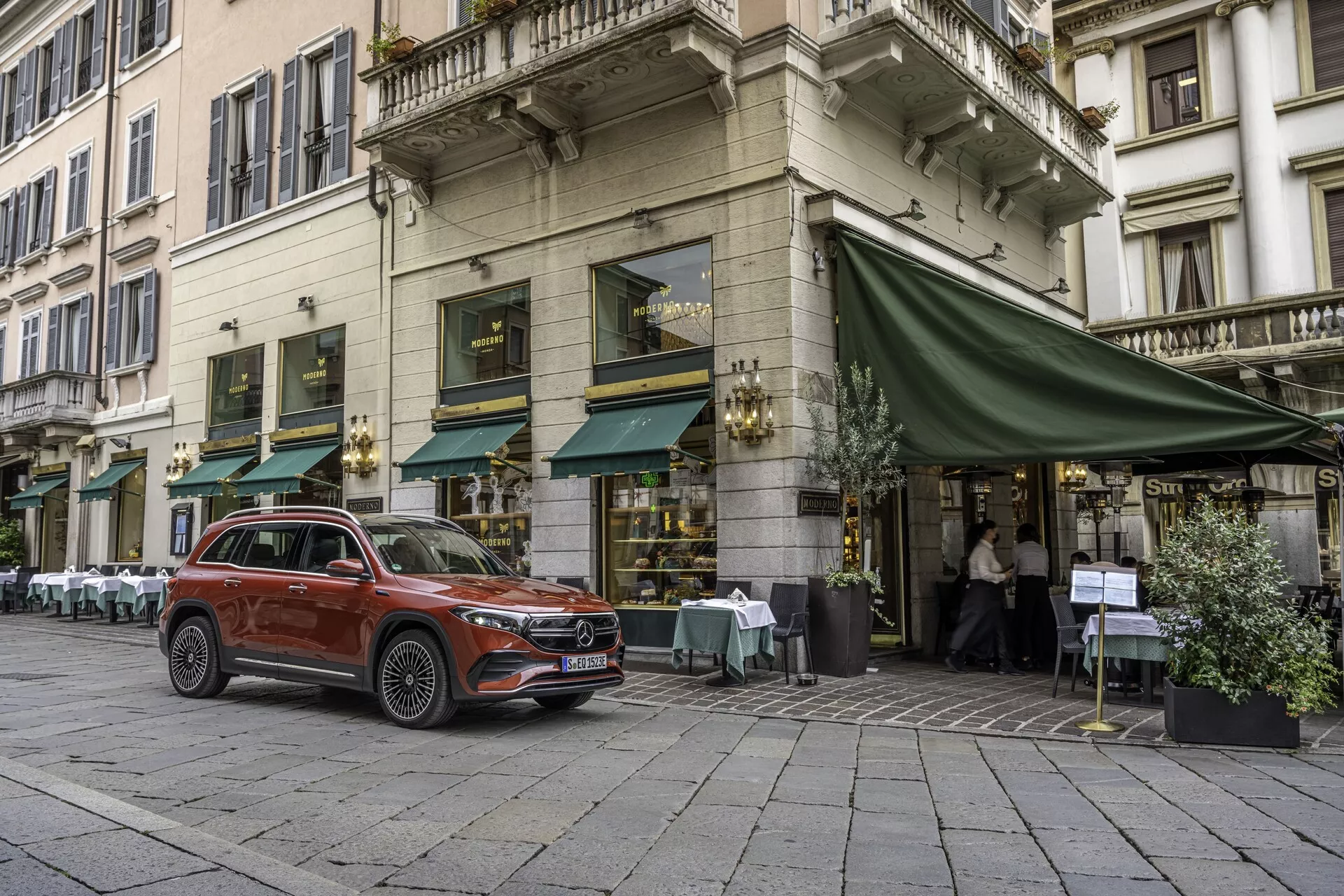 L'image capture une vue complète d'un Mercedes EQB 2021 stationné sur une rue pavée en face d'un café avec des sièges extérieurs sous un auvent vert. Photographié d'une perspective frontale trois-quarts, il montre clairement la calandre avant, les phares, le capot, la roue, la porte conducteur avant et une partie de l'arrière du véhicule. Ce SUV compact moderne présente des éléments de design tels qu'une hauteur de conduite élevée, des roues en alliage, des feux de jour à LED et une finition de peinture métallique. Son cadre urbain, probablement dans une ville européenne, met en avant son utilité en tant que véhicule familial urbain avec des caractéristiques technologiques avancées possibles, telles que des systèmes d'assistance à la conduite.
