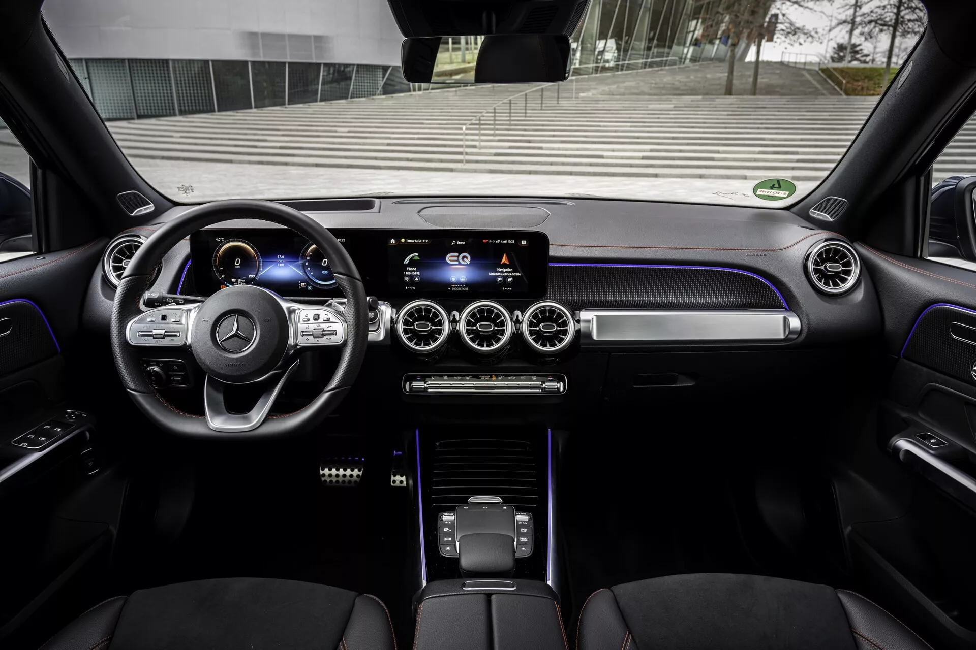 Intérieur d’un Mercedes EQB 2021 moderne vu de la perspective du conducteur. L'image offre une vue claire sur le tableau de bord, le volant à trois branches avec boutons multifonctions, le tableau de bord numérique, le système d’infodivertissement à écran tactile sur la console centrale, les bouches d'aération circulaires avec accents métalliques, les boutons de contrôle tactile de la climatisation, un sélecteur de vitesse élégant et des sièges en cuir luxueux. L'éclairage ambiant ajoute à l'atmosphère haut de gamme, avec des intérieurs qui présentent des matériaux de haute qualité et des éléments technologiques avancés.