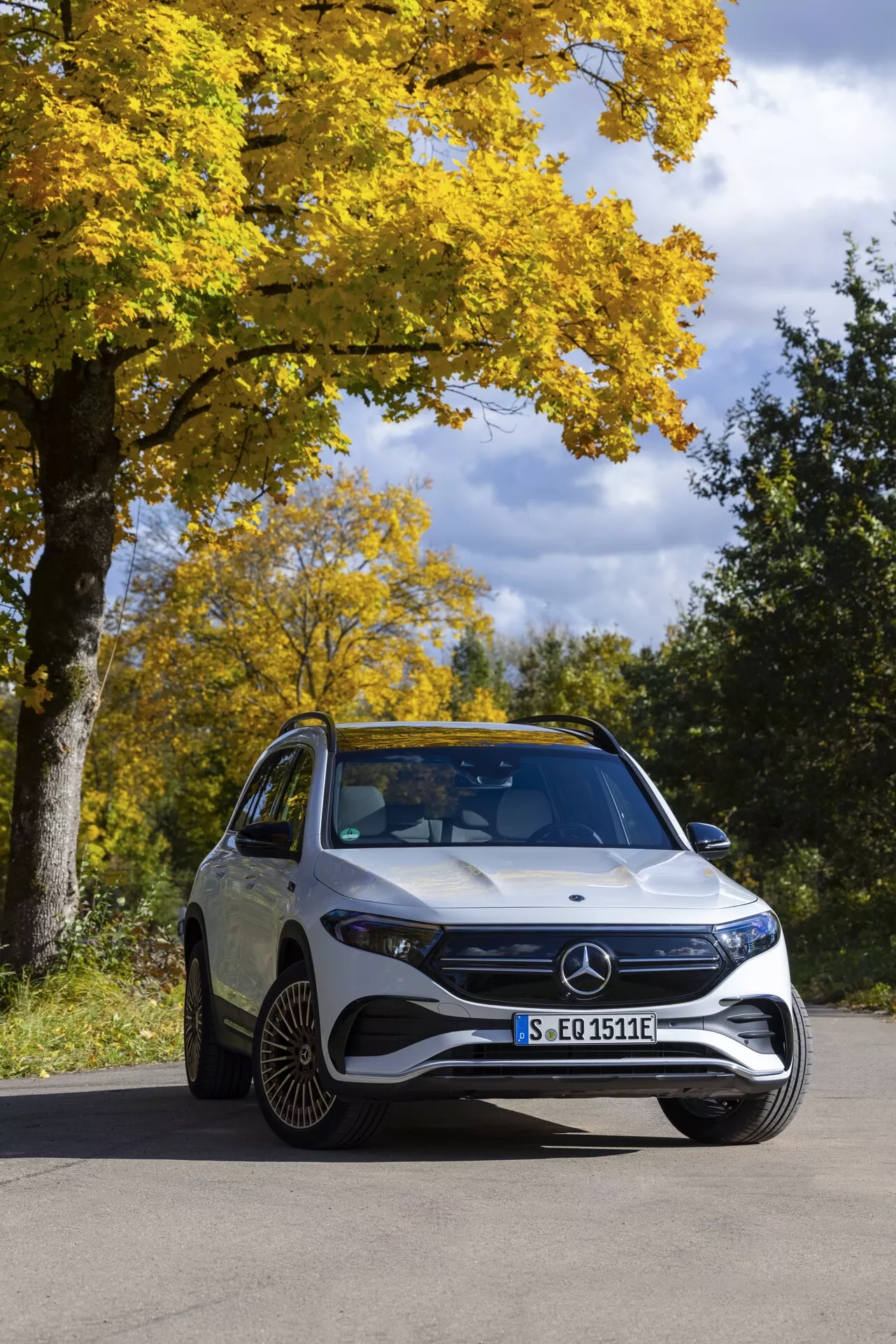 Vue de trois quarts avant d'une Mercedes EQB 2021 capturée dans un cadre naturel automnal par temps clair. Le SUV électrique de luxe présente une calandre avant proéminente avec l'emblème Mercedes, des phares LED, un pare-chocs avant sportif et des lignes caractéristiques sur le capot. Des jantes en alliage distinctives à plusieurs rayons et un pare-brise incliné sont visibles, soulignant l'efficacité aérodynamique.