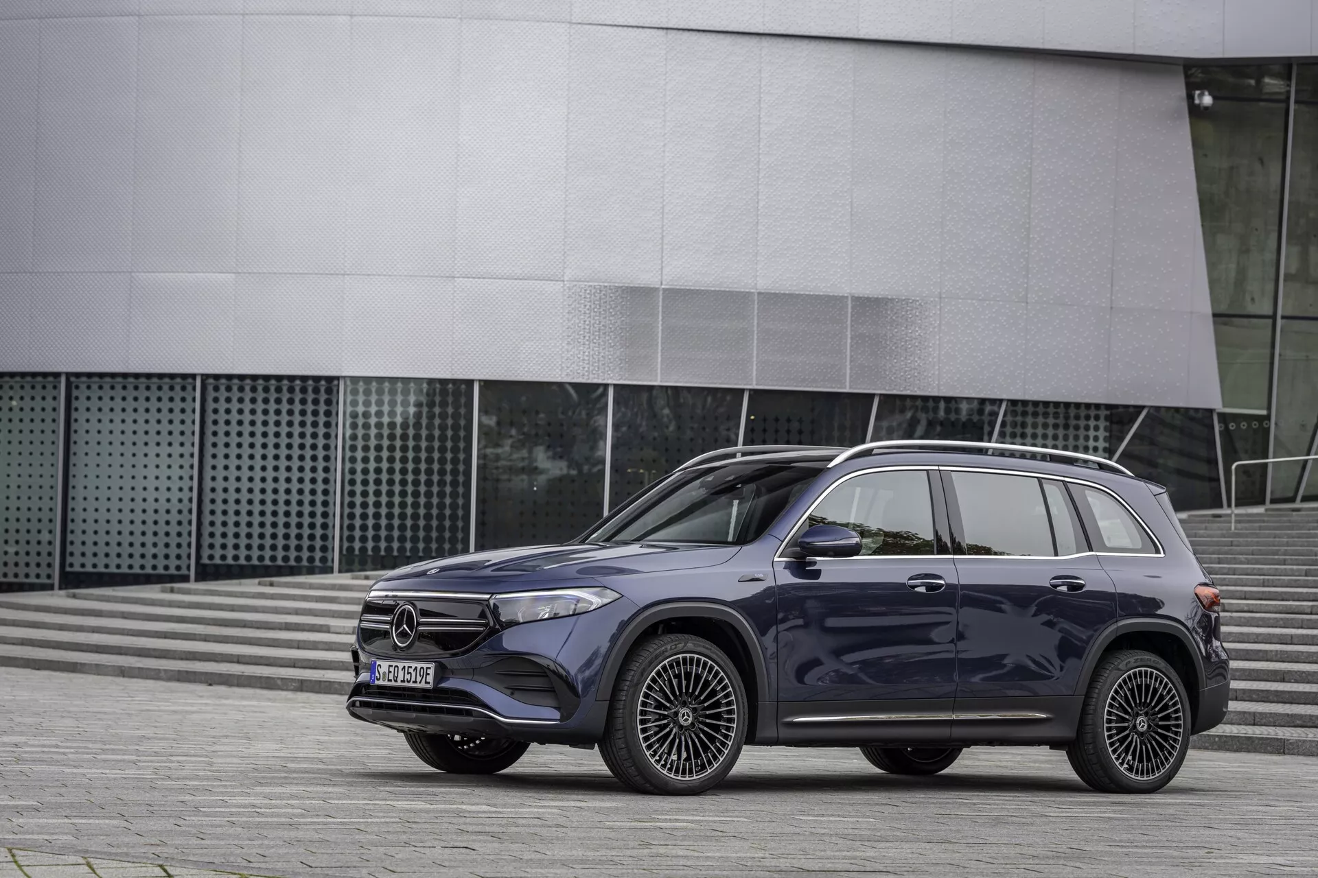 Vue de profil d'un SUV Mercedes EQB 2021 stationné dans un environnement urbain, présentant un design luxueux et élégant. Le SUV est équipé de quatre portes avec des poignées visibles, des jantes en alliage distinctives, une calandre avant proéminente avec un emblème central, des phares à LED, des rétroviseurs contrastés, des barres de toit et un toit panoramique. Les jupes latérales et les moulures des passages de roue accentuent son apparence robuste. La prise de vue en angle bas met en valeur la présence dominante du véhicule sur un fond architectural contemporain.