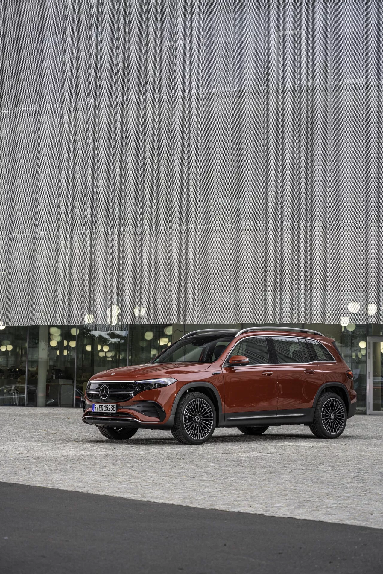 Image d'un SUV moderne Mercedes EQB (243) de 2021 de couleur rouge foncé, positionné à l'extérieur d'un bâtiment avec une façade à motif de lignes verticales. Capturé en vue de trois quarts avant, mettant en valeur la grille avant avec un emblème proéminent, des phares à LED avec feux diurnes, un capot sculpté, de grandes jantes en alliage, une ceinture de caisse haute, des entourages de fenêtre chromés et des rails de toit. Le luxe et l'utilité se mêlent dans ce design automobile contemporain, mis en valeur par un arrière-plan neutre qui met en évidence les détails de conception.
