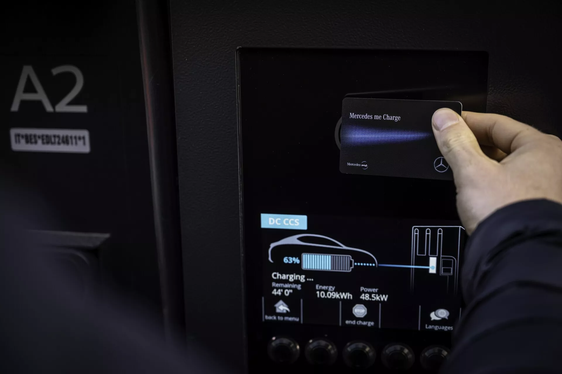 Gros plan d'une main interagissant avec l'écran tactile d'une station de charge pour véhicule électrique Mercedes EQB 2021. L'écran LCD couleur affiche 'Mercedes me Charge' en haut et indique le niveau de charge actuel à 63%, l'énergie livrée à 10,69 kWh et la puissance de sortie à 45,5 kW, avec des indicateurs graphiques de batterie et de prise. L'écran est monté sur un panneau noir mat étiqueté 'DC CCS', indiquant une charge rapide. Les détails environnants comprennent le reflet de la lumière ambiante, la texture du panneau et une étiquette 'A2' avec numéro de série à gauche.
