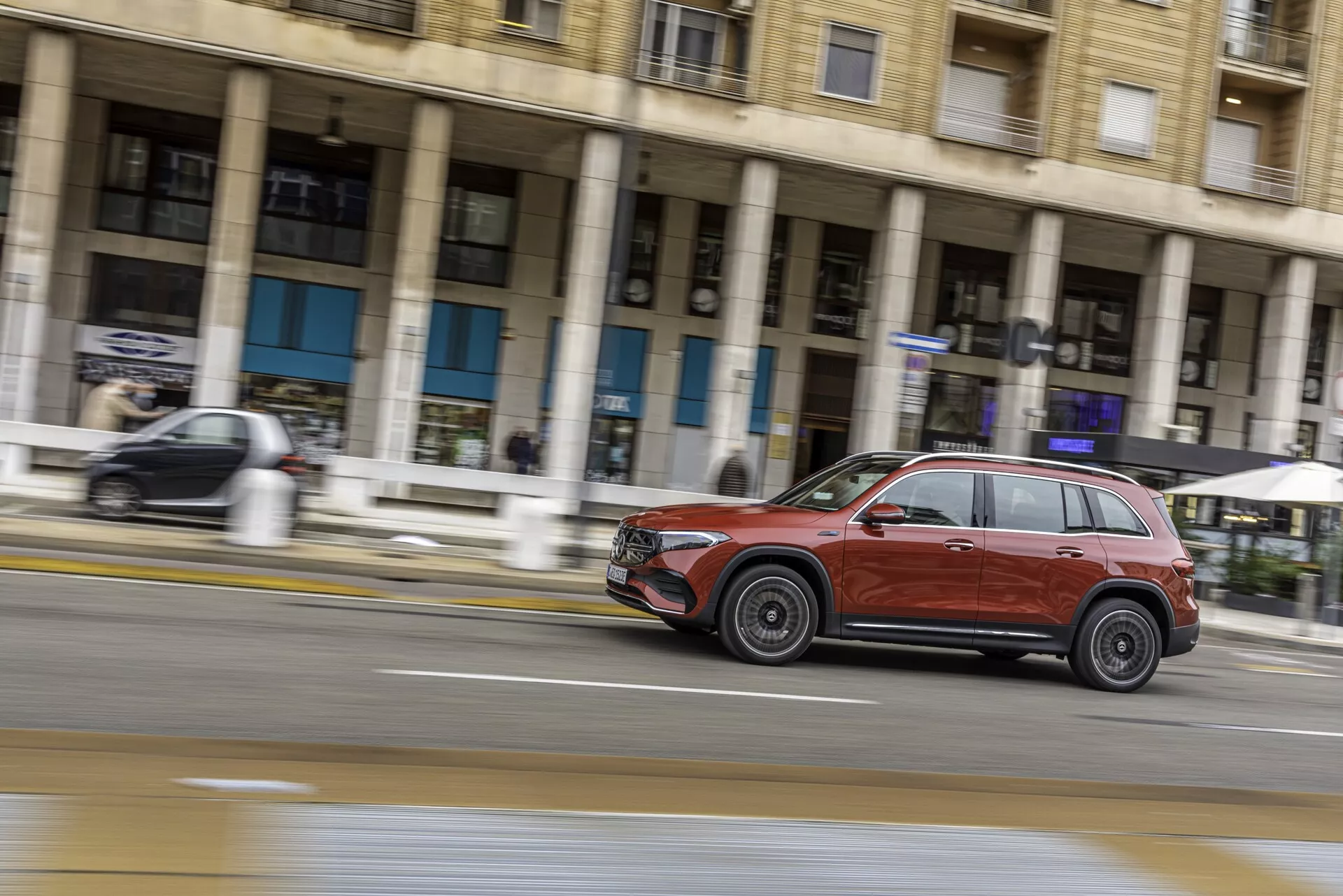Vue latérale dynamique d'un SUV Mercedes EQB 2021 de couleur rouge foncé ou marron en mouvement sur une rue de la ville. Capturée avec une technique de photographie par balayage, le profil du SUV, y compris les portes, les fenêtres, les roues, la calandre et les phares, est mis en valeur avec un flou de mouvement en arrière-plan, soulignant sa vitesse. L'image met en avant le design moderne du véhicule, les jantes en alliage et une finition brillante sous un éclairage doux et diffus.