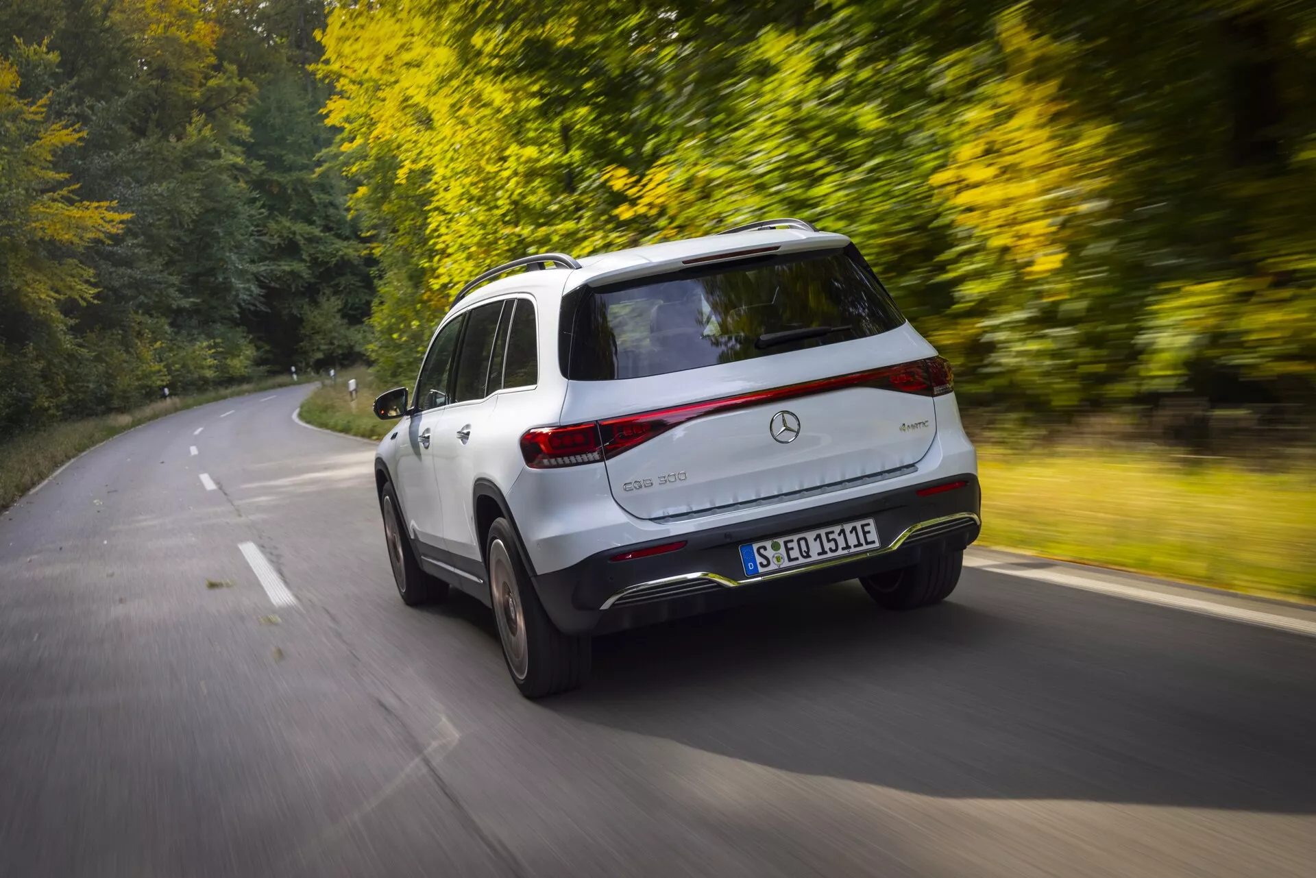 Image dynamique d'un SUV crossover Mercedes EQB 2021 en mouvement, capturée depuis une vue en trois quarts arrière surélevée. Le véhicule, majoritairement blanc, présente un design aérodynamique et élégant, mettant en valeur les profils arrière et latéraux, incluant le côté gauche, tout l'arrière, la lunette arrière, les feux arrière et une partie du pare-chocs arrière. Les aspects visibles comprennent l'emblème distinctif du véhicule, le badge du modèle et la plaque d'immatriculation. Le flou de mouvement à l'arrière-plan et aux roues suggère une conduite à grande vitesse. La route bordée d'arbres en automne et la lumière du soleil filtrant à travers les arbres ajoutent de la chaleur, présentant une expérience de conduite luxueuse et paisible, idéale pour les voyages et les aventures.