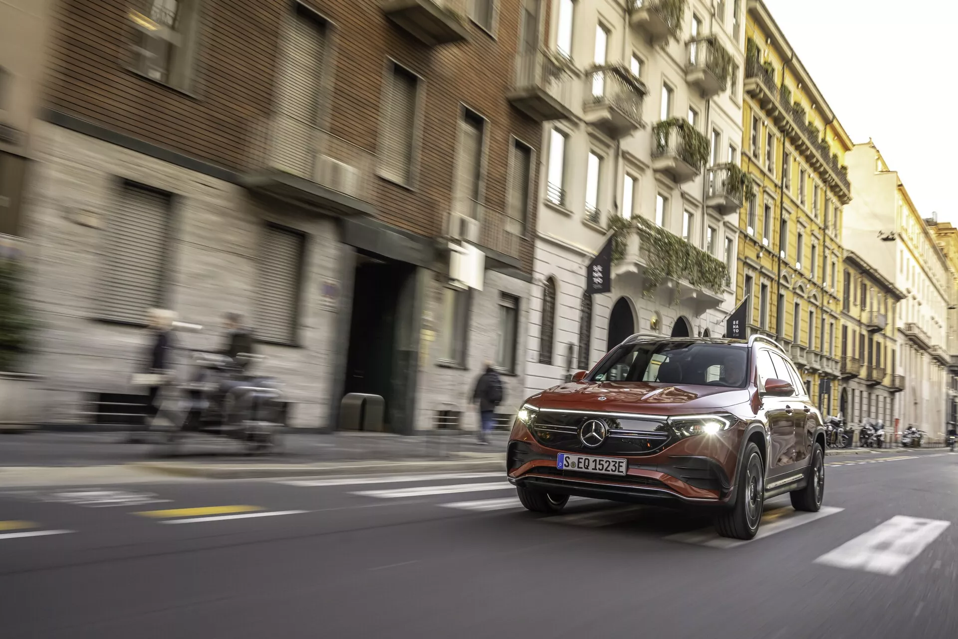 Photo en contre-plongée d'une Mercedes-Benz EQB 2021 en mouvement dans une rue urbaine, capturant la calandre avec emblème, des phares LED élégants, le pare-chocs avant, le pare-brise, une partie du profil latéral et des roues en alliage modernes. Le flou de mouvement et les traînées lumineuses créent un effet dynamique de vitesse. L'arrière-plan comprend des bâtiments historiques contrastant avec la modernité du véhicule. Cadre de jour avec des teintes chaudes et de longues ombres.