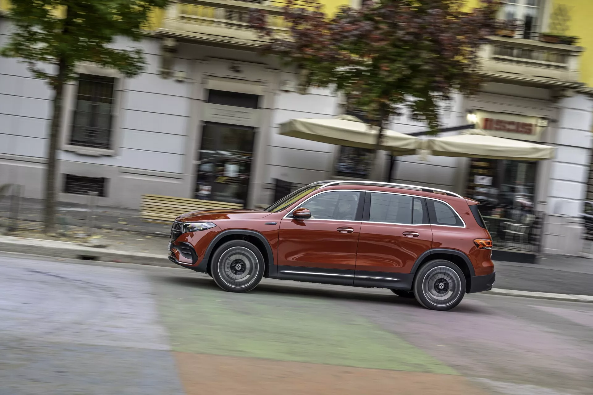 Vue latérale dynamique d'un SUV Mercedes EQB 2021 rouge métallique en mouvement, mettant en valeur ses éléments de design distinctifs tels que la grille, les phares et les jantes en alliage. Le flou en arrière-plan et sur les roues souligne sa vitesse et son agilité dans un cadre urbain.