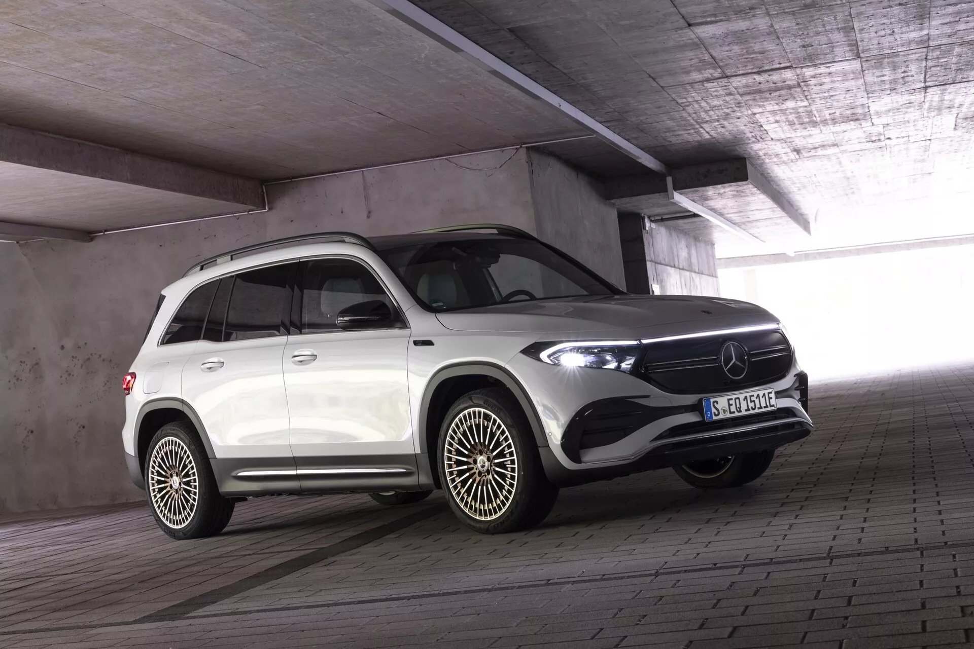 SUV de luxe blanc Mercedes EQB 2021 dans un parking intérieur avec un plafond et des piliers en béton, vue avant et latérale trois-quarts; phares LED, grille proéminente avec grand emblème et insigne électrique/hybride sur l'aile. Jantes en alliage à plusieurs branches et moulure de protection sur les portes indiquant un design tout-terrain. Le véhicule est garé avec les roues avant légèrement tournées vers la caméra, donnant une composition dynamique.
